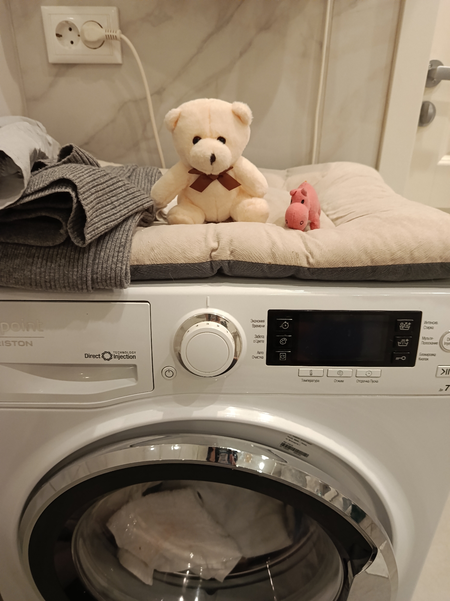 A close-up shot of a white washing machine with a gray knitted sweater, a light-colored teddy bear with a brown bow tie, and a small pink hippo toy on top of a beige cushion. The washing machine has a digital display panel with buttons and Cyrillic text. The machine's door has a chrome trim, and white clothes can be seen inside. A white electrical outlet is visible on the wall behind the washing machine. The image was taken in Odintsovo, Russia.
