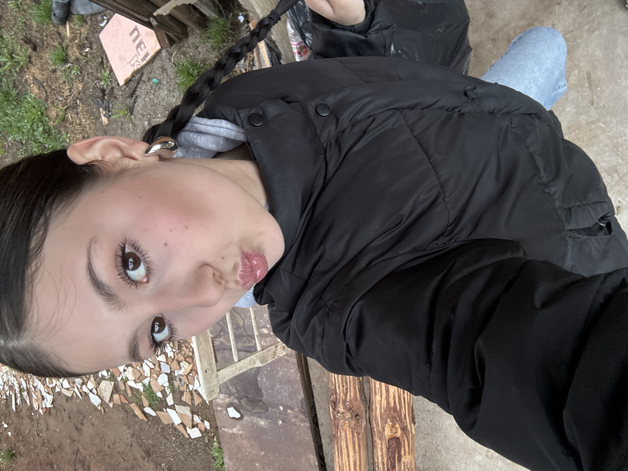 The image appears to be a selfie taken by a young woman with dark hair styled in a braid. She is wearing a black puffer jacket and a grey hooded sweatshirt. She has light skin, brown eyes, and is pouting her lips slightly. Her ear is adorned with a silver hoop earring.
The background is somewhat cluttered. There are fragments of what appears to be broken tiles or pottery scattered on the ground, mixed with dirt and some sparse patches of grass. A section of a wooden structure, possibly a fence or part of a building, is also visible. The overall environment looks rustic and somewhat untidy.
Based on the query's prompt, I can assume the location is Фоки, Russia, although no definitive landmarks are visible in the image to confirm this.