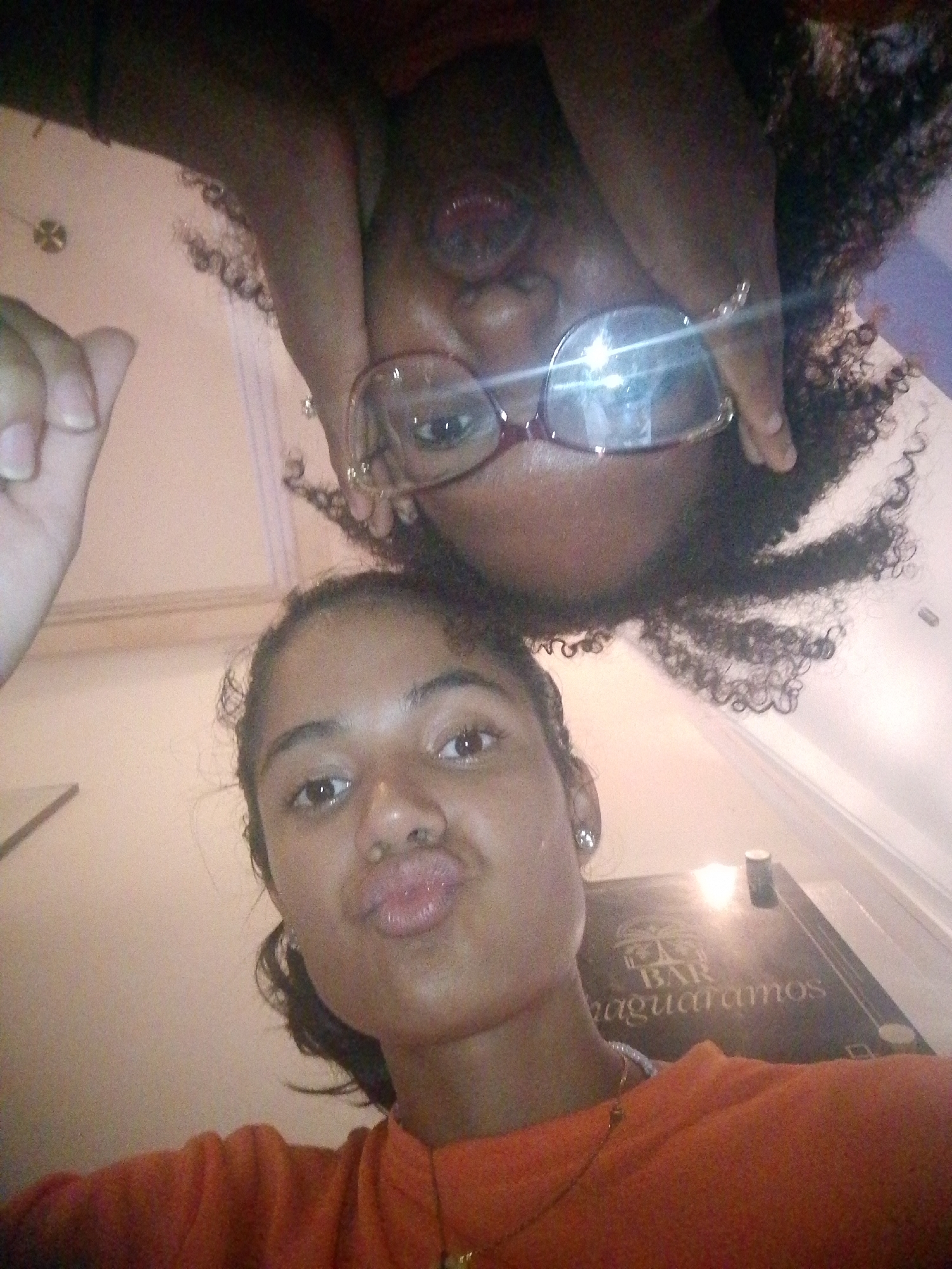 Two women pose for a selfie. One woman, closer to the camera, wears an orange top and a necklace. She purses her lips. The other woman wears glasses and has curly hair. They are in a bar in Pedro González, Venezuela, as indicated by the partially visible text "Bar naguaramos" in the background.