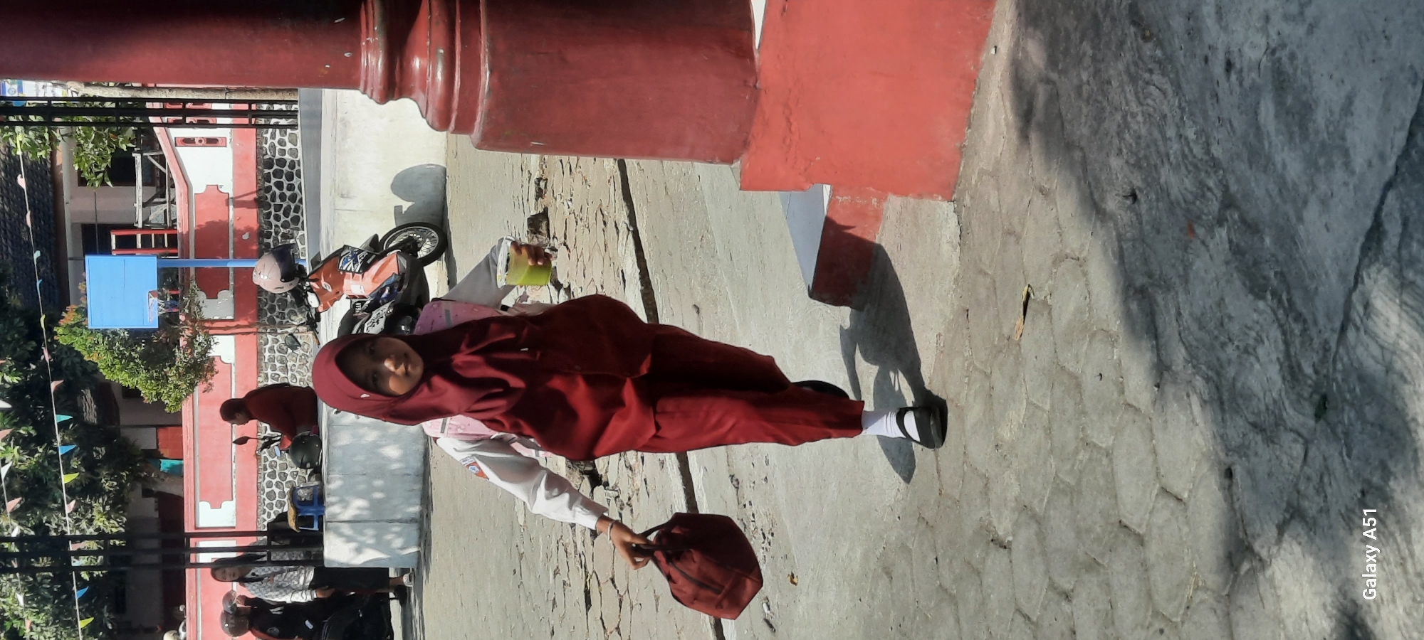 In Tangkil, Indonesia, a young girl wearing a maroon hijab and school uniform walks across a paved courtyard. She carries a maroon bag in her left hand and a clear plastic cup with a green beverage in her right. The girl is walking towards the viewer, with a school building and a basketball hoop visible in the background. A motorcycle is parked nearby. The scene appears to be set in a schoolyard on a sunny day.