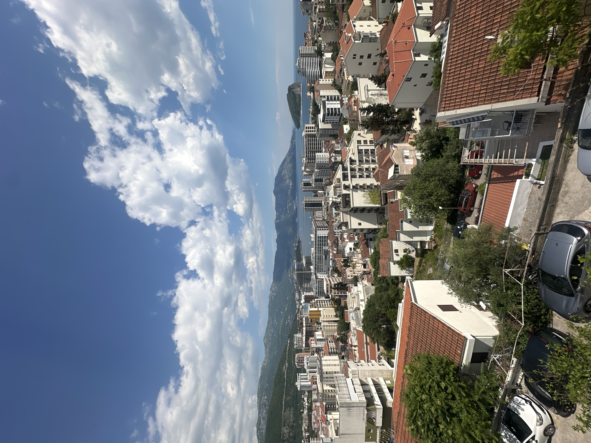 The image presents a high-angle view of Budva, Montenegro, characterized by a blend of urban and natural elements. The right side of the frame shows the tops of buildings with predominantly red-tiled roofs, interspersed with patches of greenery suggesting trees and gardens. Cars are visible, indicating residential areas and roads.
Moving towards the center, the skyline includes a mix of low-rise and high-rise buildings extending along the coast. A body of water stretches into the distance, flanked by mountains on one side.
The left side of the image is dominated by a vibrant blue sky filled with fluffy white clouds.
