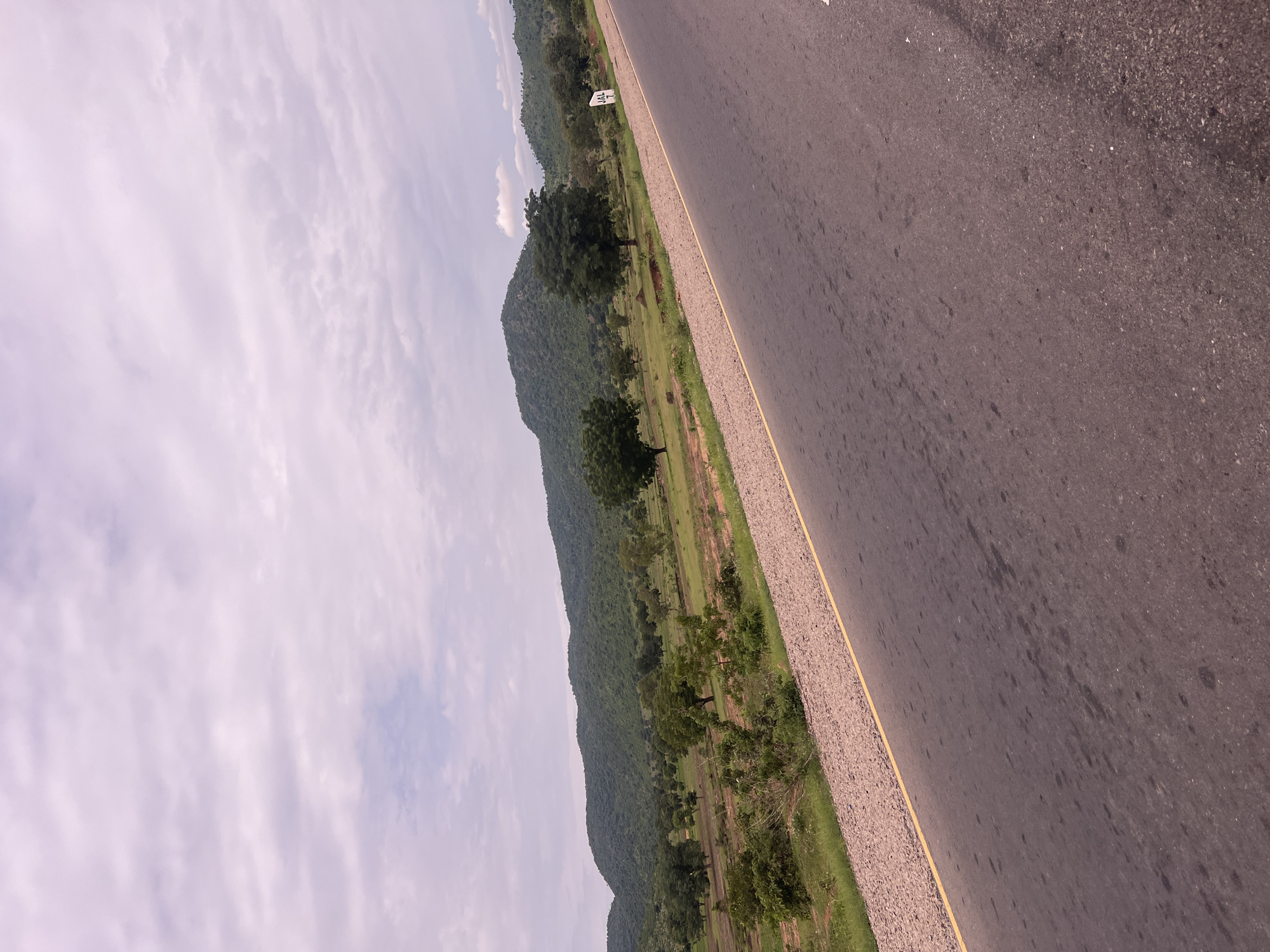 The media file captures a roadside view of a rural landscape in Minda, Nigeria. The primary subjects are a paved road stretching diagonally across the frame, a grassy embankment with scattered trees and vegetation, and a gently rolling, tree-covered hill in the background. The sky is overcast with soft, wispy clouds, suggesting a daytime scene with diffused light, possibly late afternoon.
The activity depicted is passive; it's a static view of a landscape. There's no movement or explicit event happening. The notable details include the texture of the asphalt road, the gravelly shoulder with a yellow line marking the edge, and the lush green of the natural scenery. The hill appears to be a significant geographical feature, covered in dense foliage.
A white sign with some black markings is partially visible on the left side of the road, near the embankment. While the text on the sign is not entirely legible, it appears to be a marker of some kind, possibly a distance or information post. The overall mood is serene and natural, emphasizing the expanse of the land and the open sky. The lack of any structures or people suggests a remote, undeveloped area. The image orientation is portrait, with the scene appearing tilted.
