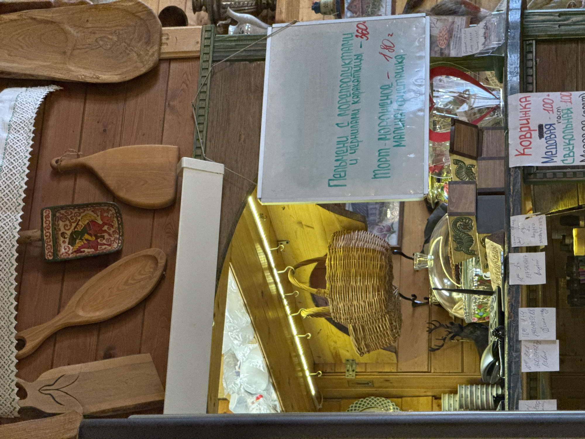 This is an indoor shot of a shop or market stall, likely in Ново-Никольское, Russia, judging by the Cyrillic script on the price signs. The primary focus is on the goods being sold.
On the left, various wooden kitchen utensils and serving ware are displayed. A doily with a lace trim is also visible.
In the center, a sign lists "Pelmeni with seafood and squid ink - 360" and "Ice cream cake - 180". A wicker basket is displayed behind the sign, and there is a glimpse of a brightly lit, white interior behind it.
On the right, another sign lists "Kovrizhka (gingerbread)" with "honey - 100" and "beet - 100". Below the sign is a variety of small items, including a glass container and a deer head decoration, as well as several pieces of paper with handwritten notes, presumably prices or inventory information. The environment appears to be a rustic, possibly traditional Russian market or shop.
