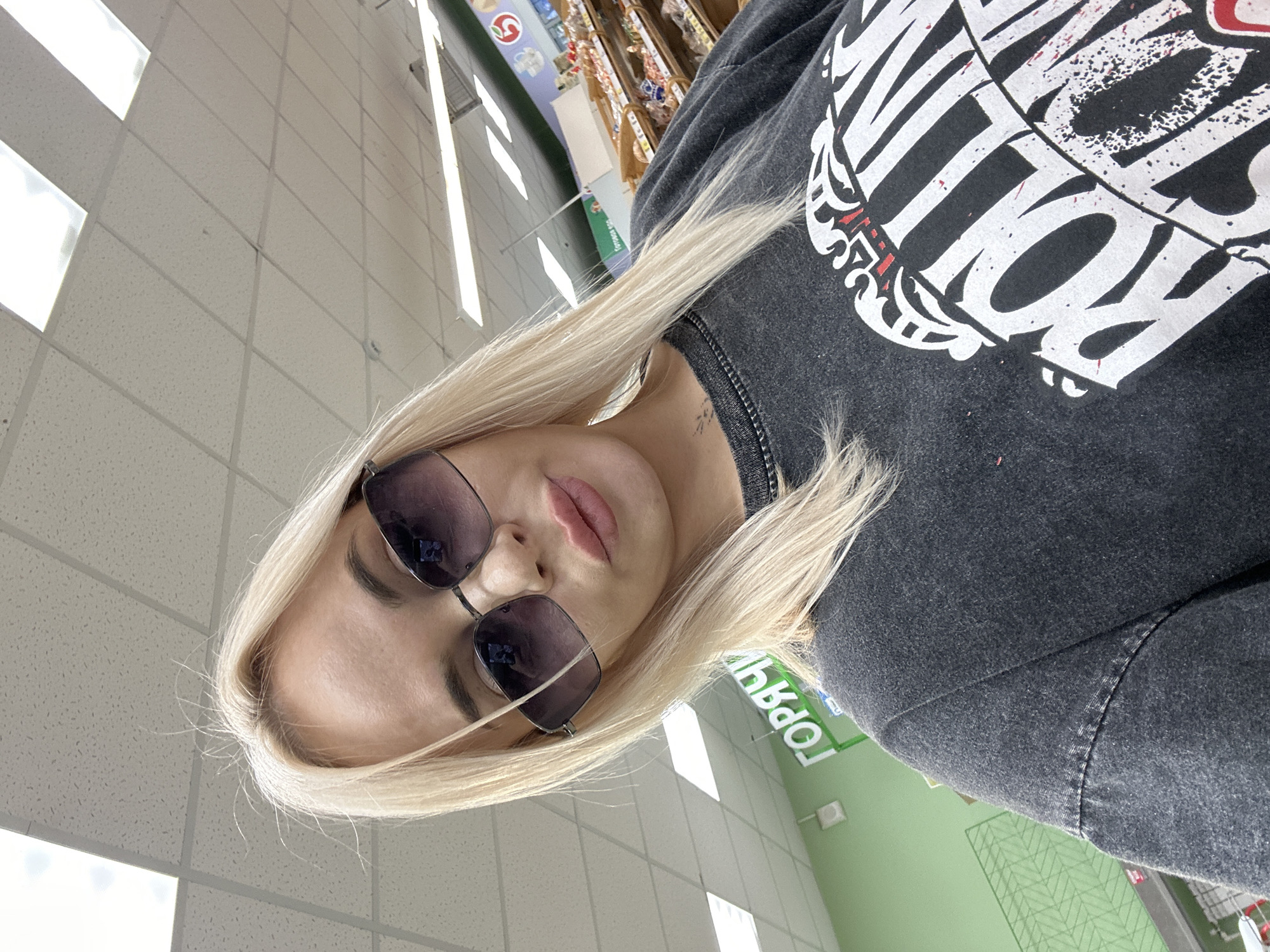 The image shows a woman with blonde hair wearing sunglasses and a dark-colored sweatshirt. The location appears to be a supermarket in Высокий, Russia, based on the Cyrillic text visible in the background.