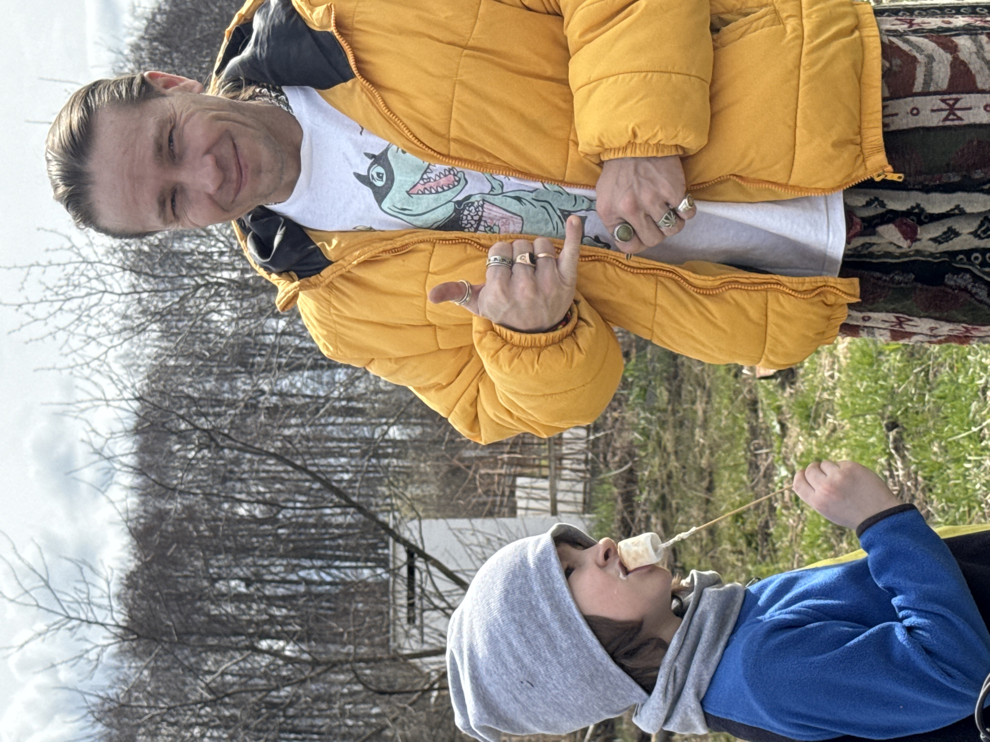 This image captures two individuals outdoors, likely in a rural or semi-rural setting, under a cloudy sky. The photograph is taken from a slightly low angle, looking upwards towards the subjects.
In the upper part of the frame, an adult male with light-colored hair pulled back is visible. He is smiling and looking directly at the viewer. He wears a bright yellow puffer jacket with a black collar and a white t-shirt underneath, featuring a prominent green cartoon dinosaur (resembling a T-Rex wearing a party hat). His left hand is raised, making a gesture with his index finger and thumb, and he wears multiple rings on his fingers. His lower body is partially obscured but he appears to be wearing patterned pants or a skirt with geometric designs.
In the lower part of the frame, a child is seen. They are wearing a grey hooded sweatshirt over a blue long-sleeved top, possibly fleece. The child is looking upwards, with a white marshmallow on a stick or string positioned at their mouth. Some white residue, likely from the marshmallow, is visible around their mouth.
The background consists of a dense area of bare trees, suggesting late winter or early spring. Patches of green grass are visible on the ground in the lower right, indicating the onset of warmer weather. A faint structure, possibly a small building or fence, can be seen amidst the trees behind the child.
The overall impression is one of a casual outdoor activity, possibly involving snacks or a simple outing in Noviy, Russia. The adult appears cheerful, while the child is focused on their treat. No explicit text is visible in the image, aside from the graphic on the adult's t-shirt.