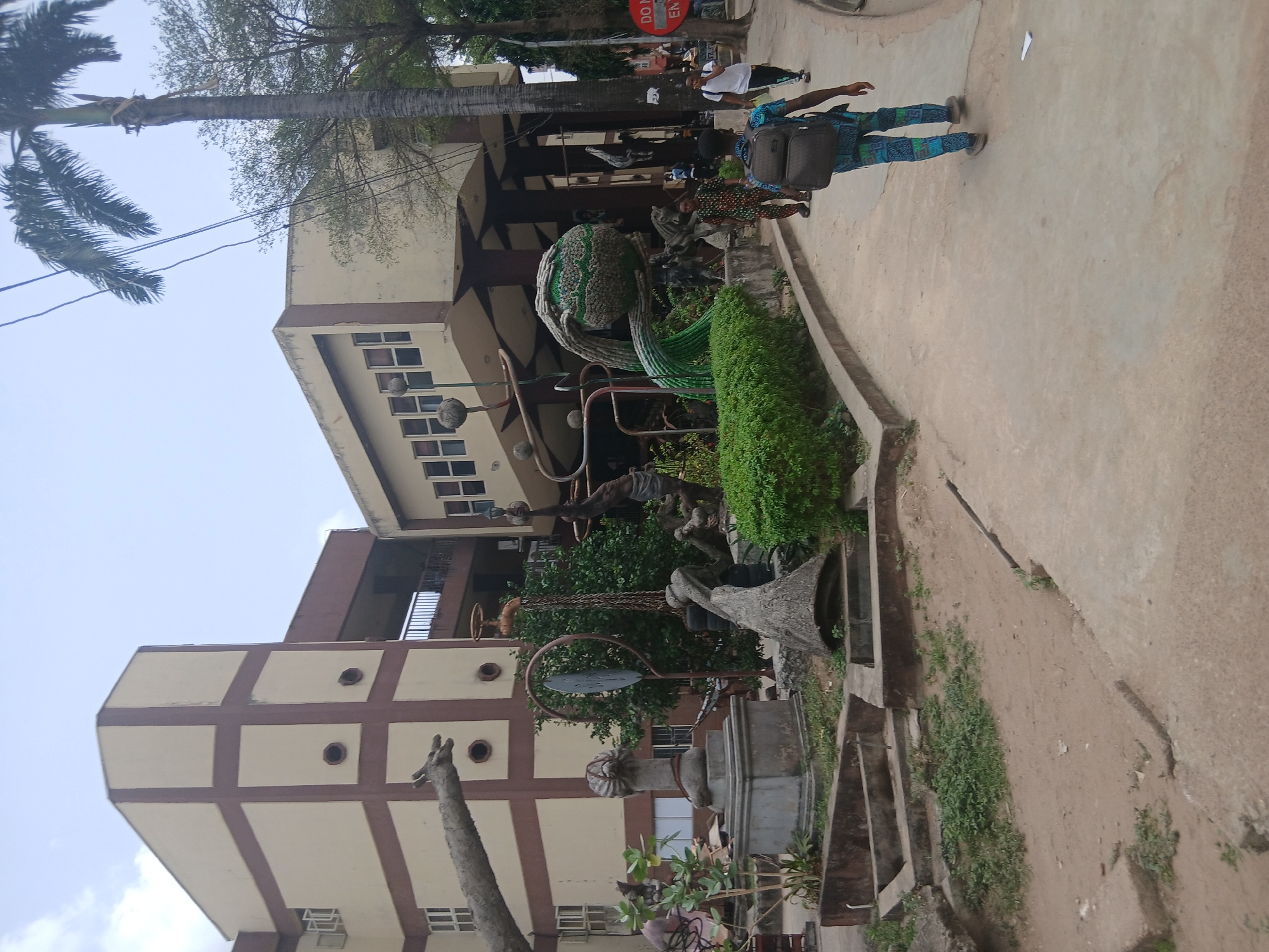 The image shows a daytime scene in Lagos, Nigeria. A multi-story building with a tan facade and brown grid pattern is visible on the left. In front of the building is a public space with sculptures and landscaping. One sculpture is a globe-like structure with green tendrils. Other sculptures made of metal and concrete are also present. A paved walkway cuts through the scene from the bottom right to the top, where a few individuals are walking. One person is wearing a patterned green and blue outfit and carrying a backpack. A "Do Not Enter" sign is partially visible in the background. There are trees providing shade.