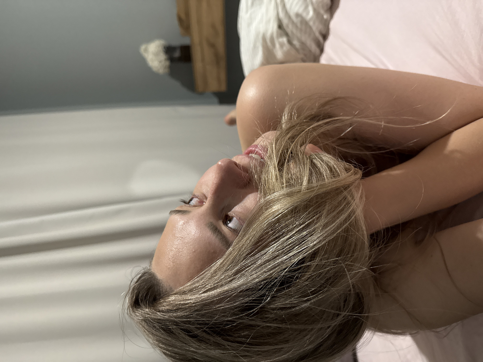 The image features a woman in a close-up, medium shot, oriented sideways. Her head is positioned on the right side of the frame, looking upwards towards the top edge. She has long, blonde or light brown hair that cascades across her shoulders and towards the bottom of the frame. Her eyes are open, gazing slightly upwards, and her lips are subtly parted, hinting at a gentle smile. Her expression appears relaxed and contemplative. Part of her bare arm is visible, raised and bent, with her hand likely near her head, partially obscured by her hair.
The setting appears to be indoors, likely a bedroom. In the blurred background on the left, a light grey or bluish-grey wall is visible, with faint horizontal lines that could be part of wall paneling or a headboard. Further to the left and top, a wooden object, possibly a shelf or furniture element, and a fluffy white decorative item are vaguely discernible. On the right side of the frame, behind the woman's head and arm, there's a soft pink surface, likely a bedsheet or blanket. The lighting is soft and even, suggesting indoor artificial light or diffused natural light. There is no visible text in the image. The overall mood conveys a sense of quietude and intimacy.