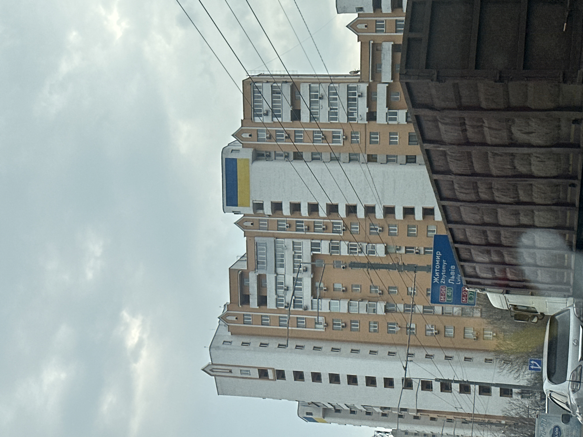 The image shows a tall apartment building in Kyiv, Ukraine, partially obscured by a vehicle on the right. The building is predominantly white with light brown accents. A blue and yellow Ukrainian flag is visible near the top. A street sign for Zhytomyr and Lviv is present. Overhead are power lines. The sky is overcast. Other vehicles are in the foreground.