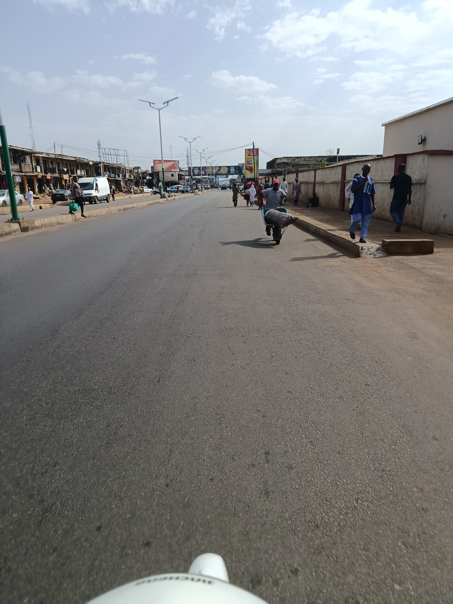 The image depicts a street scene in Jos, Nigeria. The road is asphalted and appears to be a main thoroughfare. On the left side of the road, there is a row of shops and buildings with open fronts. Several people are walking along the sidewalk in front of these establishments, and some vehicles, including a white van, are parked or moving on the road. Street lights are visible along the roadside.
On the right side of the road, there is a concrete wall with a building behind it. Several people are walking on the sidewalk and along the edge of the road. One individual is pushing a wheelbarrow loaded with sacks. The sky is cloudy, and the overall impression is of a busy urban environment. The perspective of the photo suggests it was taken from a moving vehicle or a person on a bicycle or motorcycle, as the bottom of the image shows a part of a white object, possibly a helmet or vehicle bodywork.