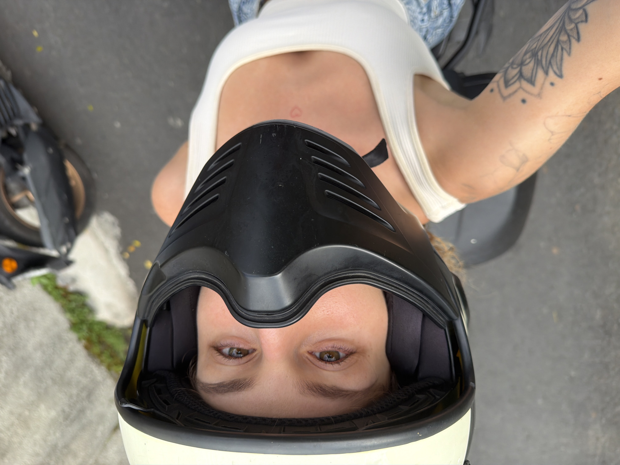 A first-person perspective shot shows a person wearing a beige and black motorcycle helmet, with their head tilted back. The person's face is visible, with brown eyes and eyebrows. They are wearing a white tank top, and a portion of their right arm is visible, showing a tattoo. The background appears to be a street with asphalt, and a portion of a black motorcycle is visible to the right. The image does not provide any identifiable landmarks or information that would allow for precise location determination within North Kuta, Indonesia.