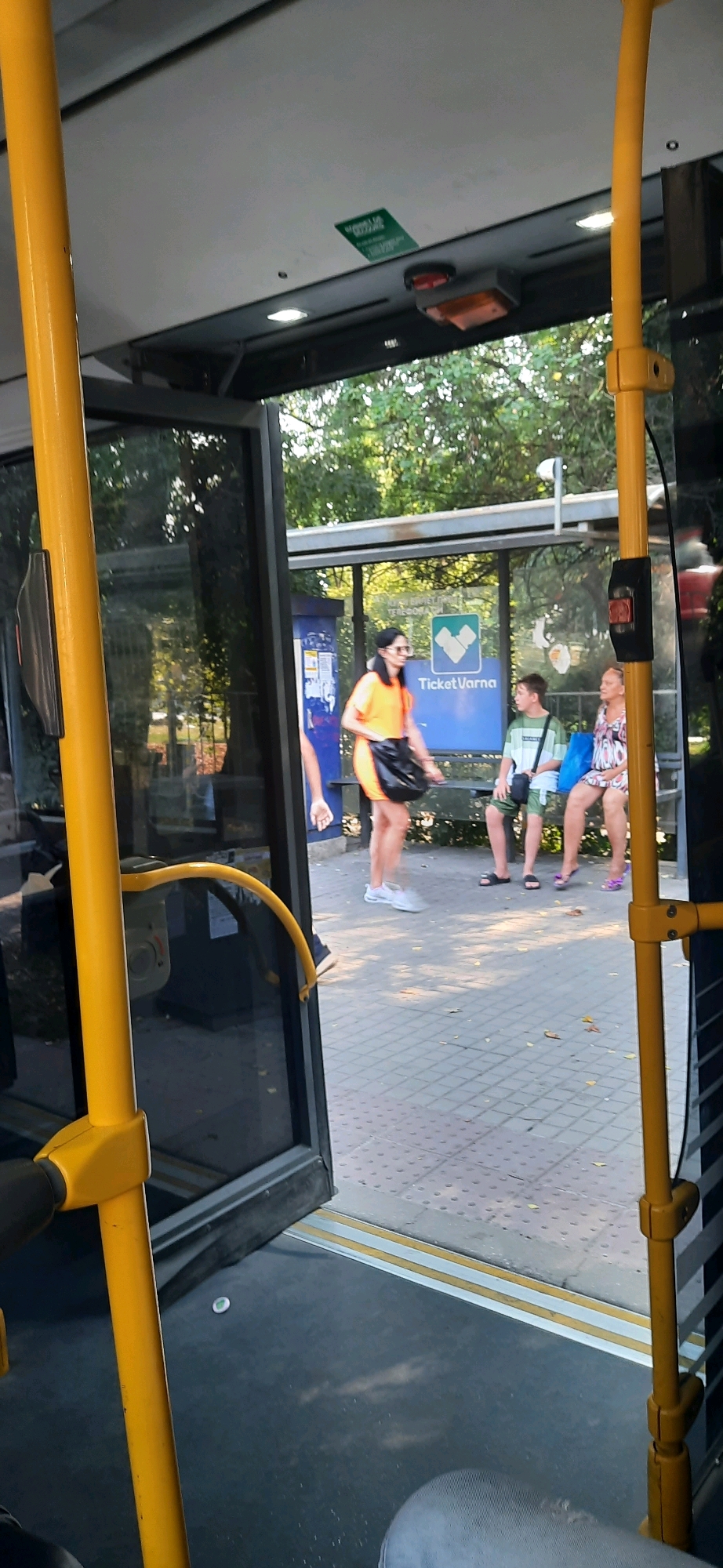 The image depicts a view from inside a bus, looking out towards a bus stop in Varna, Bulgaria. A yellow pole is prominently visible on the left side of the frame, with a yellow handrail curving towards the right. Through the open bus doors and windows, a bus stop shelter with a blue sign featuring the "Ticket Varna" logo is visible. Three individuals are present at the bus stop. A woman in an orange dress stands near the front, holding a black bag, while a young boy and an older woman sit on a bench. The ground at the bus stop is paved with grey square tiles. Green foliage and trees surround the bus stop. The overall lighting suggests it is daytime.