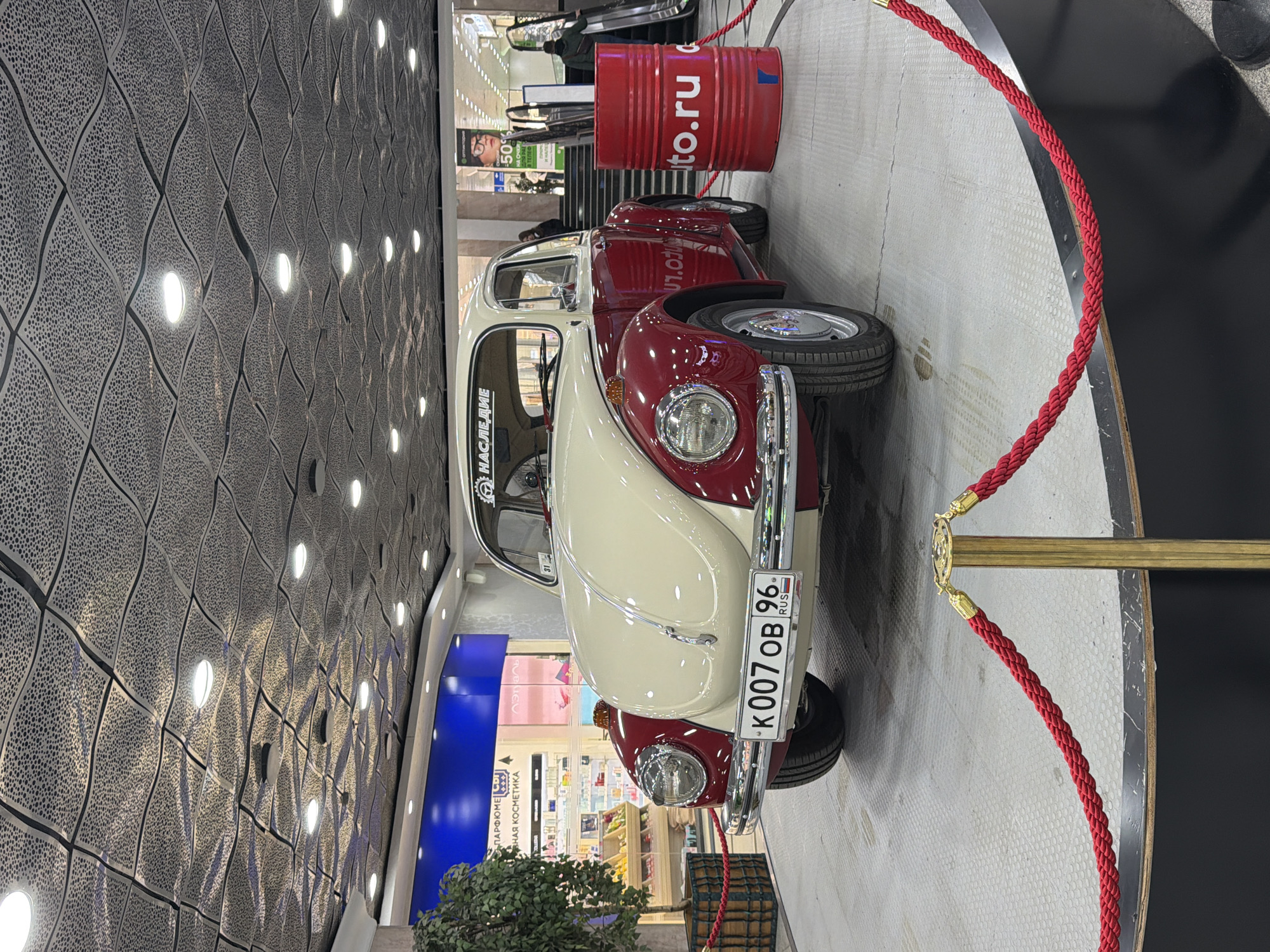 The image captures a vintage two-tone Volkswagen Beetle displayed inside a modern shopping mall in Yekaterinburg, Russia. The car, painted cream on the upper body and maroon/dark red on the lower half, is situated on a circular platform and enclosed by red velvet ropes with brass stanchions, indicating it is an exhibit.
A red barrel, marked with "auto.ru," stands behind the car on the platform. The car's license plate, "K007 OB 96 RUS," is prominently visible, with "96" being the regional code for Sverdlovsk Oblast. A sticker on the rear windshield reads "НАСЛЕДИЕ" (NASLEDIE), meaning "Heritage."
The background reveals a bustling mall environment. To the left, a decorative wall features a dark, patterned surface with numerous recessed circular lights. To the right, escalators are visible, with a few blurry individuals on them, ascending to other levels. Retail storefronts line the mall interior; one sign clearly displays "Л'Этуаль" (L'Etoile) along with "ПАРФЮМЕРИЯ И КОСМЕТИКА" (Perfumes and Cosmetics). Another sign advertises "50% НА ОЧКИ" (50% off glasses), with the word "ОПТИКА" (Optics) also partially visible. The scene is brightly lit by artificial light, characteristic of an indoor commercial setting, suggesting either daytime or evening. No specific weather conditions are applicable. The overall impression is a public display of the classic car within a shopping center.