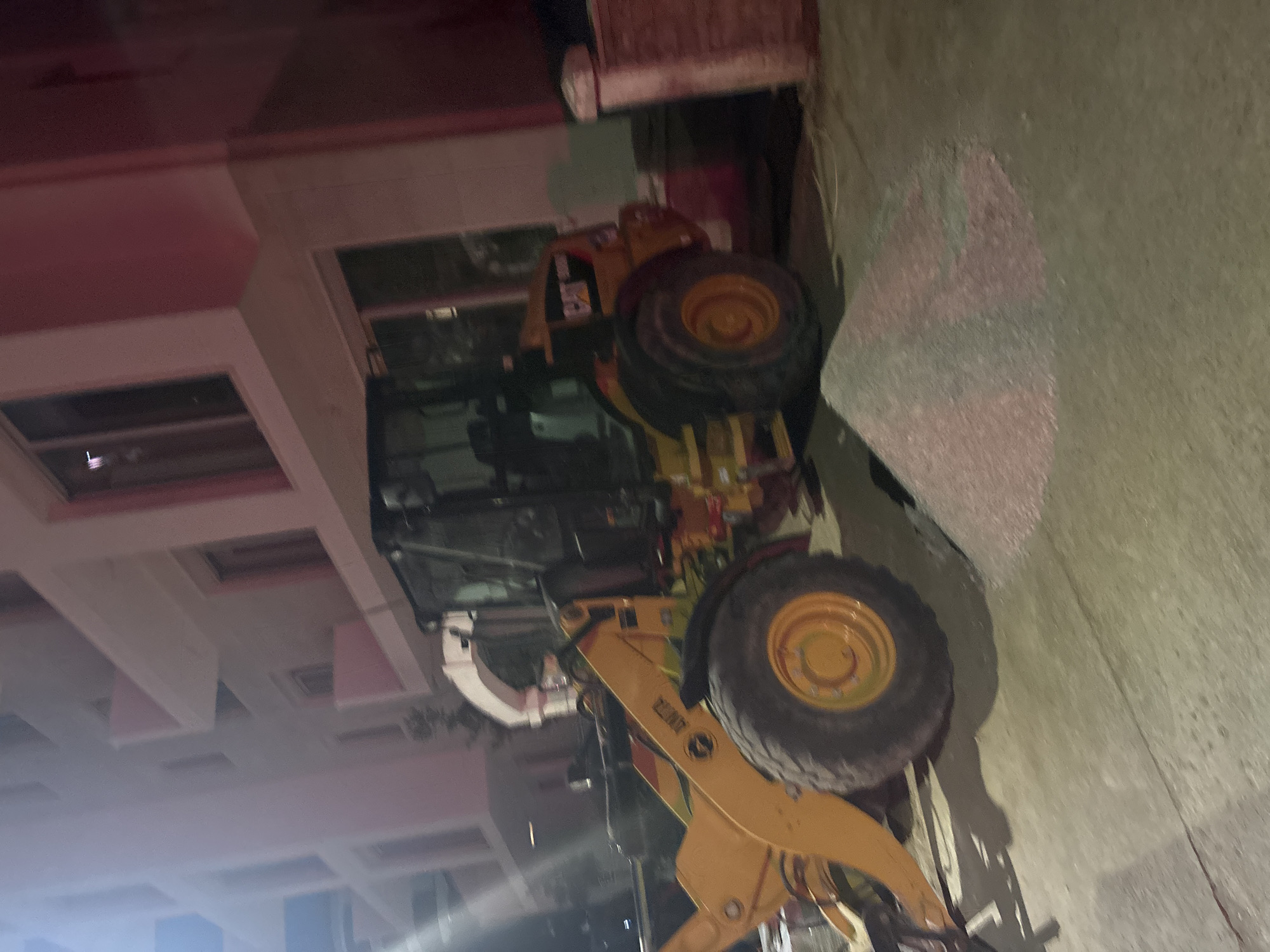 An outdoor scene at night shows a yellow construction vehicle, possibly a backhoe or loader, parked in front of a multi-story building. The building appears to be a modern hotel or apartment complex with balconies and large windows. A pile of aggregate material is on the ground in front of the vehicle. The scene is dimly lit, with some artificial lighting casting shadows. The location is identified as Budva, Montenegro.