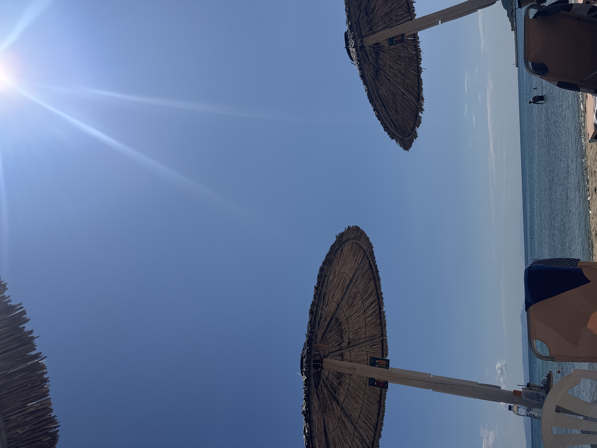 This image captures a sun-drenched scene at a beach in Stavros, Greece. Two thatched umbrellas provide shade over beach chairs, with a glimpse of the clear blue sea and distant hills. The sun is bright, casting a glare in the upper right corner. The water is calm, and a few people are visible in the distance, some wading in the sea. Beach chairs are arranged on the sand. The overall impression is one of a peaceful, sunny day at a coastal resort.