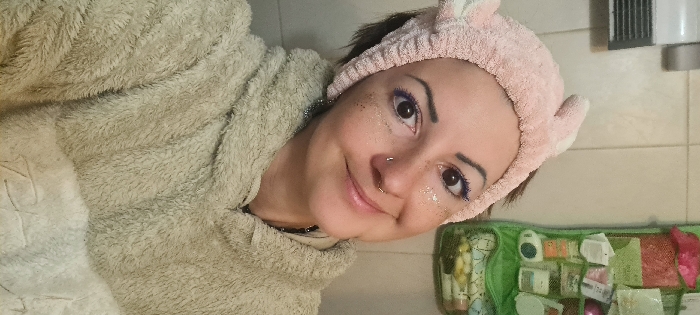 A female subject, appearing to be an adult, is depicted in a close-up, waist-up portrait within a bathroom in Nahariyya, Israel. The subject has dark hair covered by a pink terry cloth headband with ear-like protrusions. Her face features brown eyes with purple eyeliner and glitter applied under the eyes and on the cheeks. She has a gold-toned hoop septum piercing and a small stud nostril piercing. Her lips are glossy, and she exhibits a slight smile. She is wearing a light beige or off-white fuzzy hooded garment. In the background, a tiled wall is visible. To the upper left, a white wall-mounted electric heater is present. To the right, a green hanging mesh organizer filled with various toiletries and personal care items is mounted on the wall.