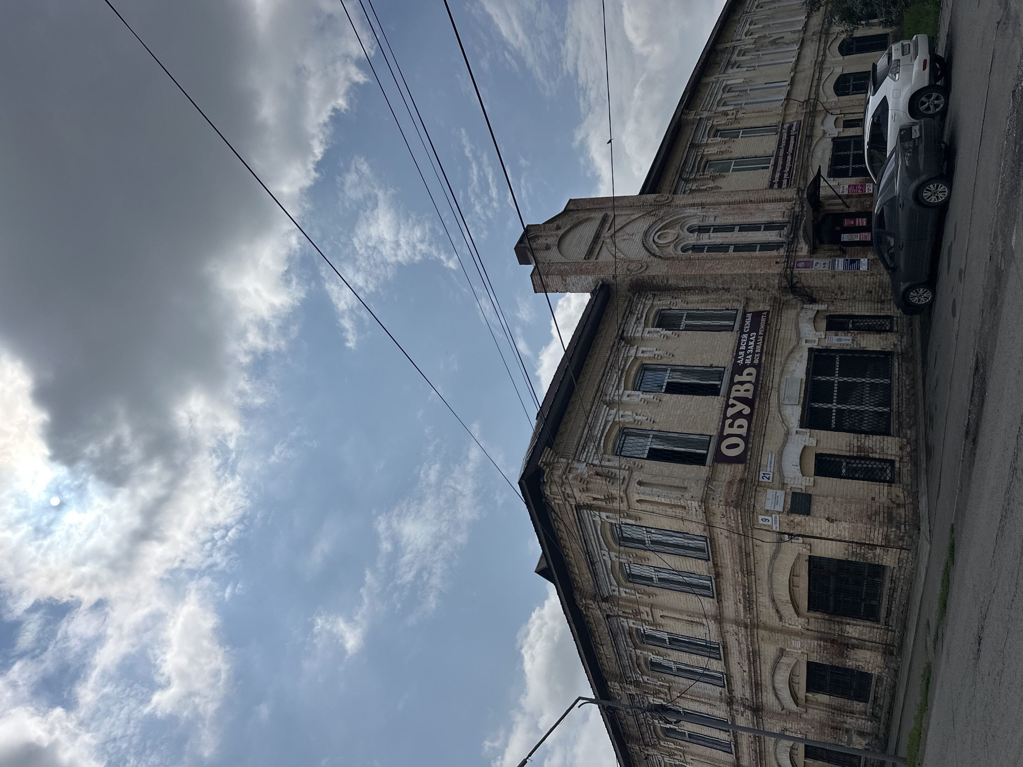 The image captures an exterior view of a weathered, yellow brick building in Nogornyi, Russia, under a partially cloudy sky. The building exhibits architectural details such as ornate window frames and decorative brickwork. A sign above a storefront reads "ОБУВЬ" (Shoes) in large letters, with smaller text below indicating services like "ALL FOR THE FAMILY - FOR ORDER - ALL TYPES OF REPAIR." Several windows on the ground floor are covered with metal grates. Two cars, a dark sedan and a white SUV, are parked on the street in front of the building. Overhead, several power lines are visible against the sky. The environment appears to be an urban street scene.