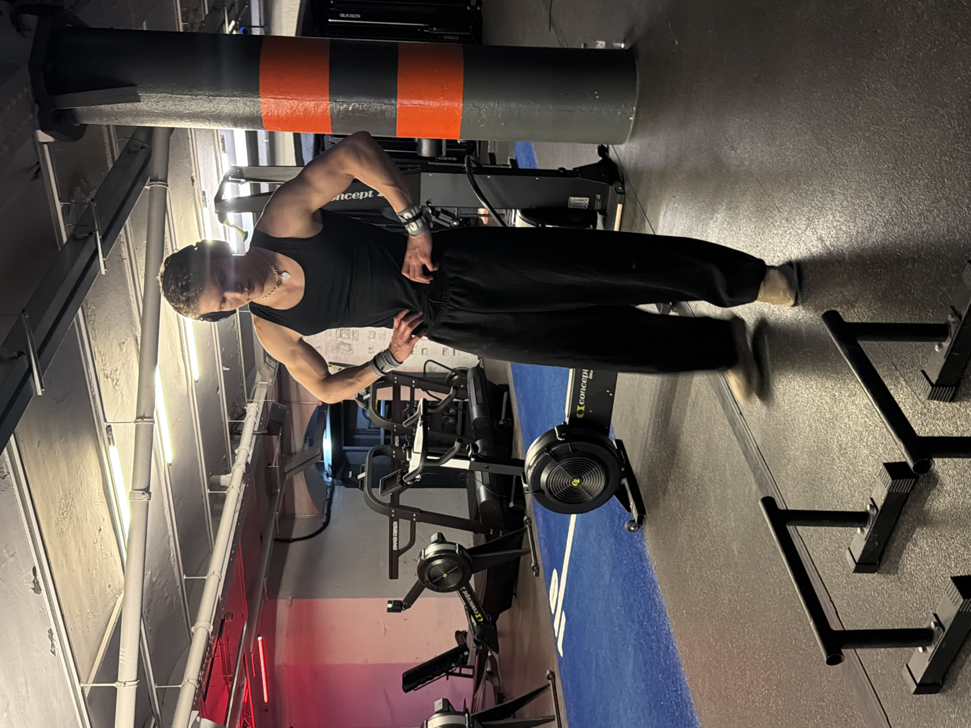 The image depicts a person standing in what appears to be a gym. The person is wearing a black tank top and black pants, with wristbands on both wrists, and a necklace. They are standing with their hands on their hips. Behind the person are several rowing machines, and to their right are pull-up bars. There is a blue line on the floor next to the rowing machines. A large pipe, painted with orange and grey stripes, runs along the ceiling. The lighting is somewhat dim and artificial. The specific location in London is not discernible from the visual information provided.