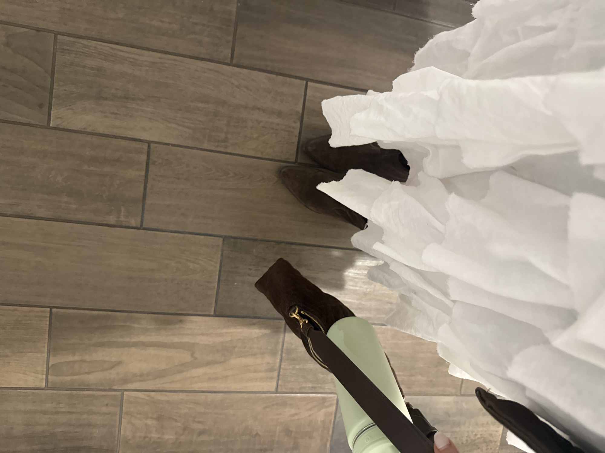 A top-down view shows a person standing on a tiled floor, presumably indoors. The flooring consists of long, rectangular tiles arranged in a running bond pattern. The person is wearing brown boots. A large quantity of white tissue paper is visible, extending upwards, obscuring parts of the scene. A light green bottle with a brown strap is visible. No specific location is discernible.