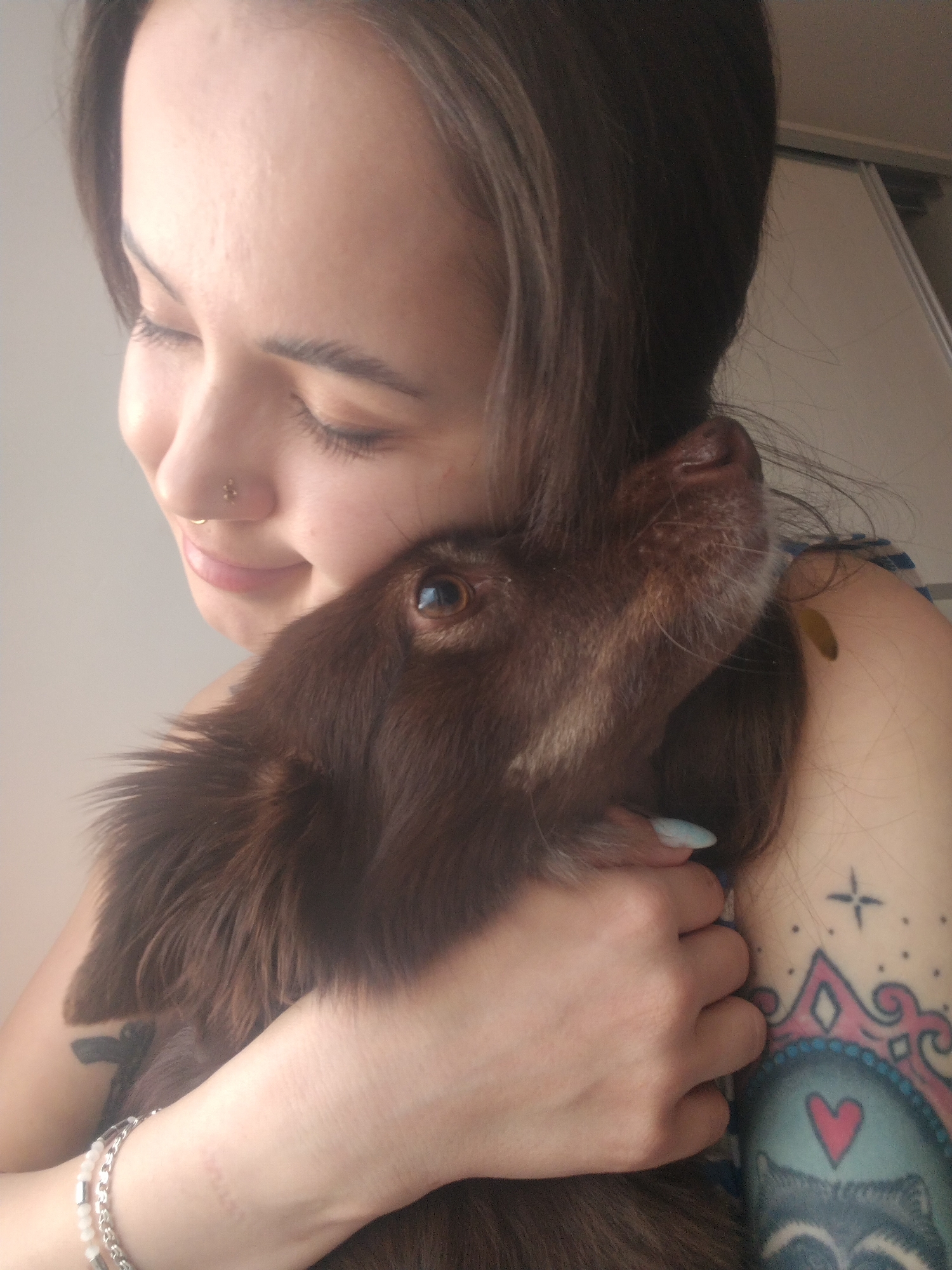 The image captures a warm and intimate indoor scene featuring a young woman embracing a brown dog. The woman, with dark hair cascading around her shoulder, is seen from the upper torso, smiling gently with her eyes closed, conveying a sense of peace and affection. She wears a gold-colored stud and hoop nose ring. Her left arm, adorned with a beaded and silver-chain bracelet, gently holds the dog. Visible on her left shoulder is an elaborate tattoo featuring a large heart within a stylized animal face, possibly a racoon, framed by decorative elements in shades of pink and blue, along with a simpler black tattoo on her bicep. Her fingernails are painted a light blue.
The brown-furred dog is nestled closely against her, its head resting near her neck, with one dark, reflective eye looking upwards. The dog's expression and posture indicate comfort and a strong bond with the woman. The background is a plain, light-colored wall, possibly part of a cabinet or wardrobe, suggesting a private, domestic setting, likely during daytime hours. The overall mood of the image is one of mutual love, contentment, and companionship. This heartfelt moment takes place in Noviy, Russia. No visible text is present in the image.