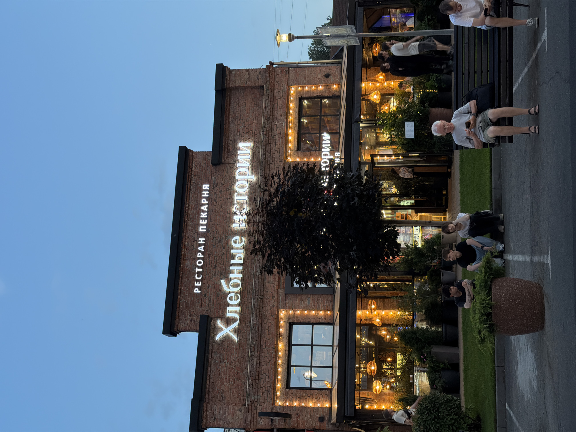The image shows a restaurant and bakery named "Хлебные истории" (Khlebnye istorii, meaning "Bread Stories") located in the city of Krasnodar, Russia. The building is constructed of brick and features illuminated signage. Several people are visible in the scene: three young men are sitting on the grass in the foreground, while an older man and another man are seated on a bench in the middle ground. Other individuals are seen walking or standing near the entrance of the establishment, which is decorated with string lights and features large windows revealing the interior with pendant lighting. The scene appears to be set during dusk, with the sky a deep blue.