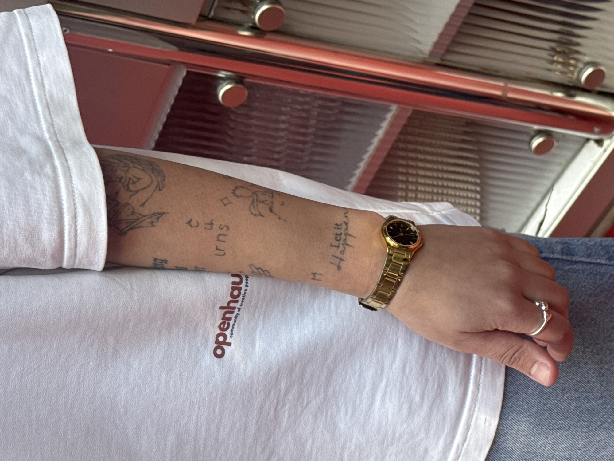 A close-up shot shows a person's arm and hand, with the arm adorned with tattoos and a gold wristwatch. The tattoos appear to be minimalist in design, featuring small figures, symbols, and lettering. The wrist watch has a black dial. The hand is wearing a silver ring. The person is wearing a white t-shirt with a logo "openhau community of creative people" on the lower left side and blue jeans. The background is a blurred view of the interior of a building or vehicle.