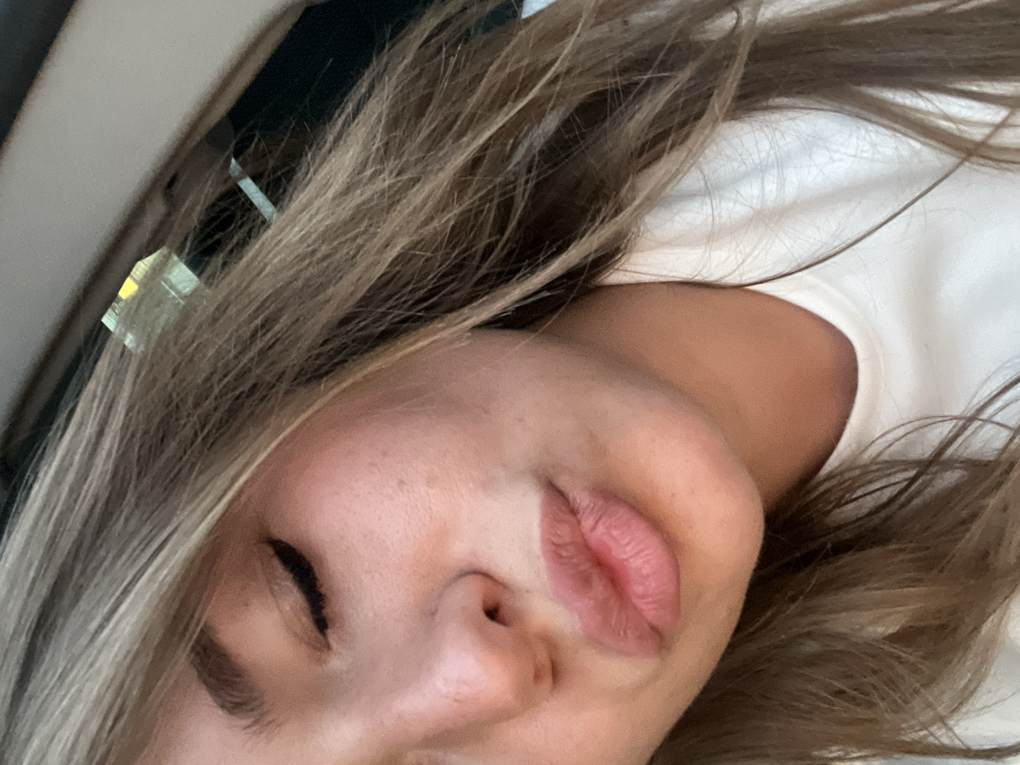 A close-up, angled shot of a person's face, likely female, with blonde and brown hair, lying on their side. The person's eyes are closed, and the shot focuses on their facial features, including their nose and lips. The background suggests an indoor setting, possibly a car interior or a room, with glimpses of white and neutral tones. There is nothing in the image to indicate a specific location, particularly Budva, Montenegro.