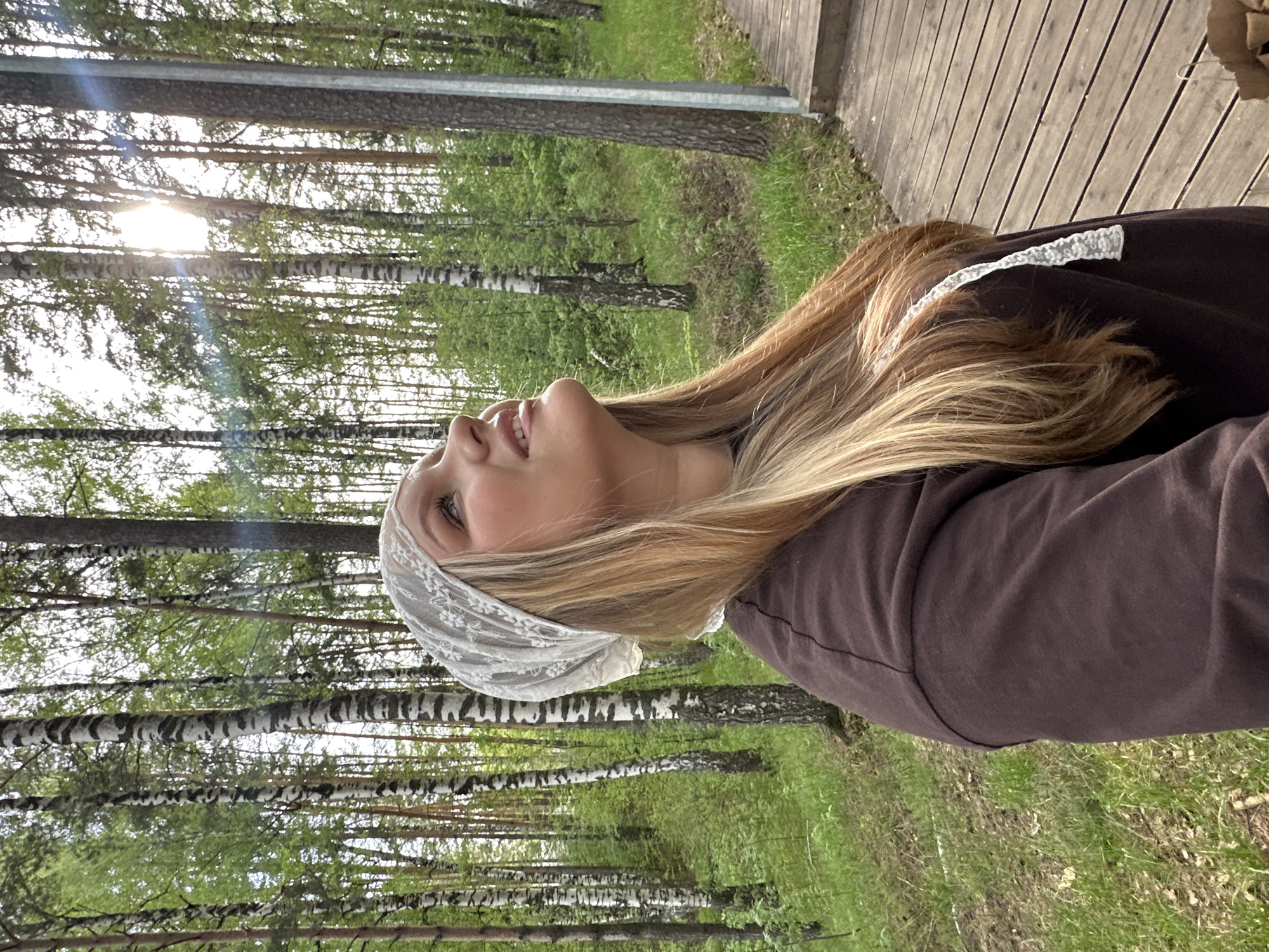 A young woman with blonde hair and wearing a white lace head covering and a brown sweatshirt is in a forest. The woman is looking up with a slight smile. White birch trees are visible in the background. Part of a wooden boardwalk or walkway is visible on the right. The location is likely in the city of Chaikovsky, Russia.