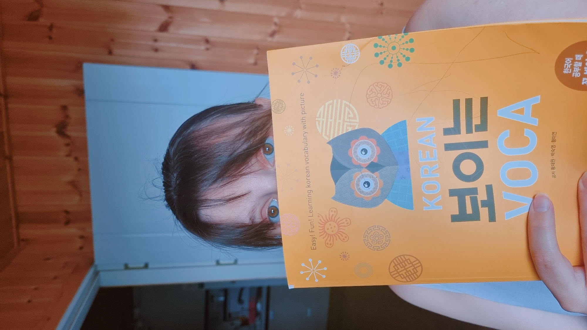 A woman is holding an orange "Korean Voca" vocabulary book in front of her face, only her eyes and the top of her head are visible above the book. The book has images of an owl and various Korean symbols, along with text in Korean and English. The background consists of a wooden wall on the left and a light blue wall on the right. The location may be in Dunnae-myeon, South Korea.