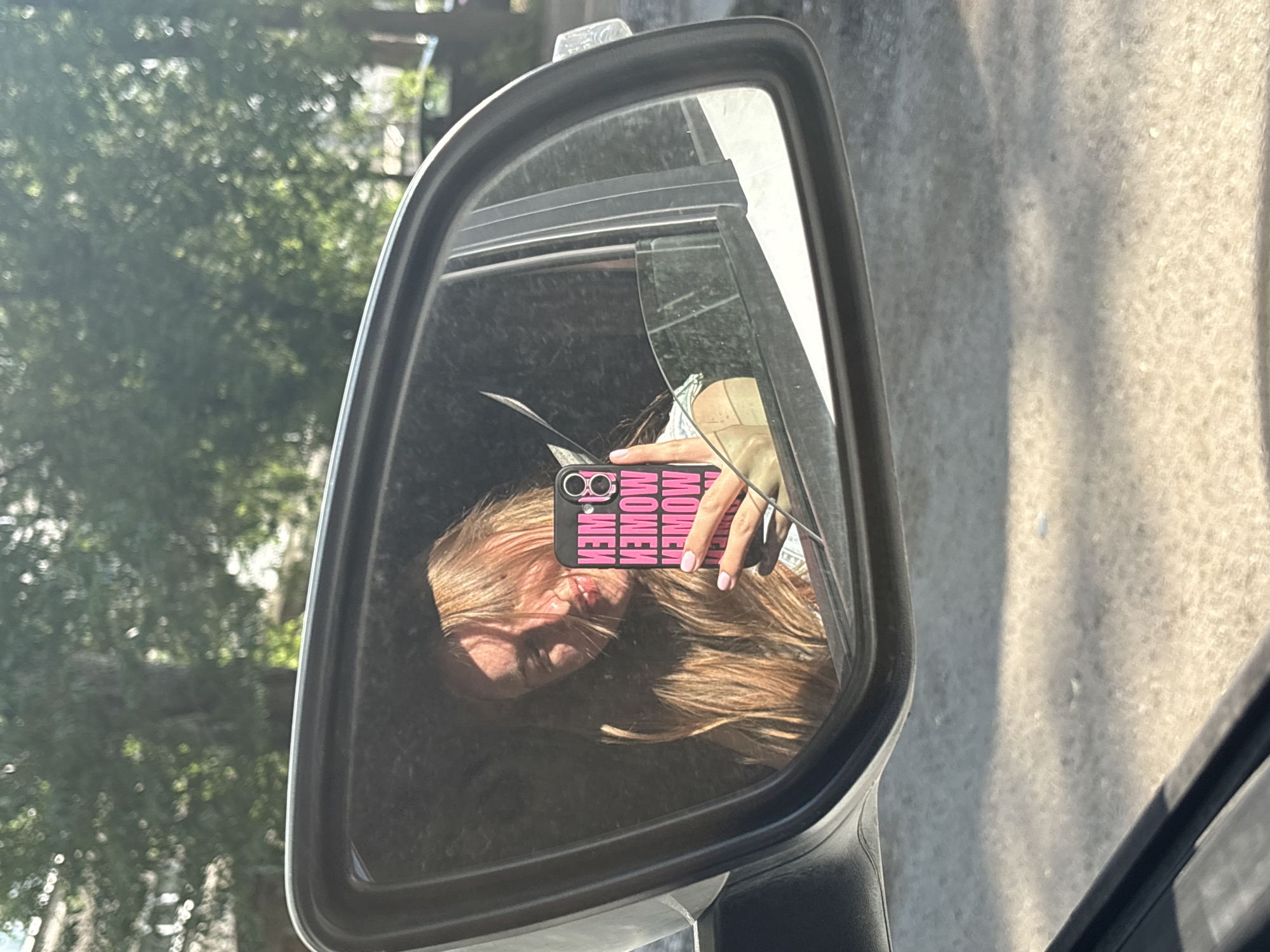 The image shows the reflection of a woman in a car's side mirror. She is holding a cell phone with a pink case that has the word "WOMEN" repeatedly printed on it. Part of her face and her hand holding the phone are visible in the mirror, along with some of her blonde hair. The mirror is reflecting the inside of the car and an obstructed view of a tree outside. The photo was not taken in Perm, Russia; this information cannot be extracted from the image.