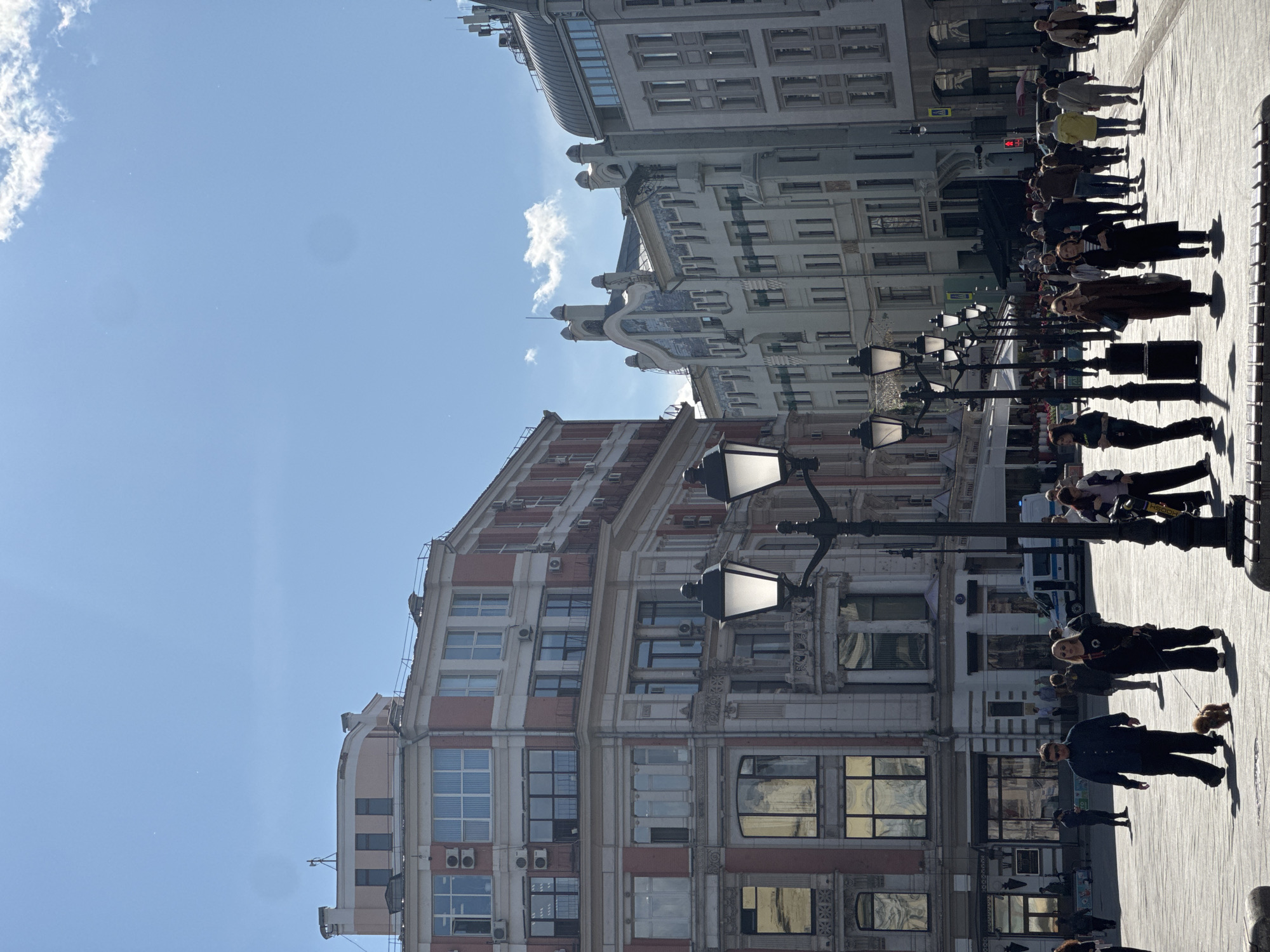 This image shows a street scene in Moscow, Russia, on a sunny day. Buildings with elaborate architectural details line both sides of the street, featuring a mix of red and white facades. The street is bustling with pedestrians, who are walking in both directions. Several lampposts stand along the street, adding to the traditional urban atmosphere. The sky is clear and blue with a few scattered clouds visible. The perspective is a vertical shot, giving a clear view of the buildings and the activity on the street.