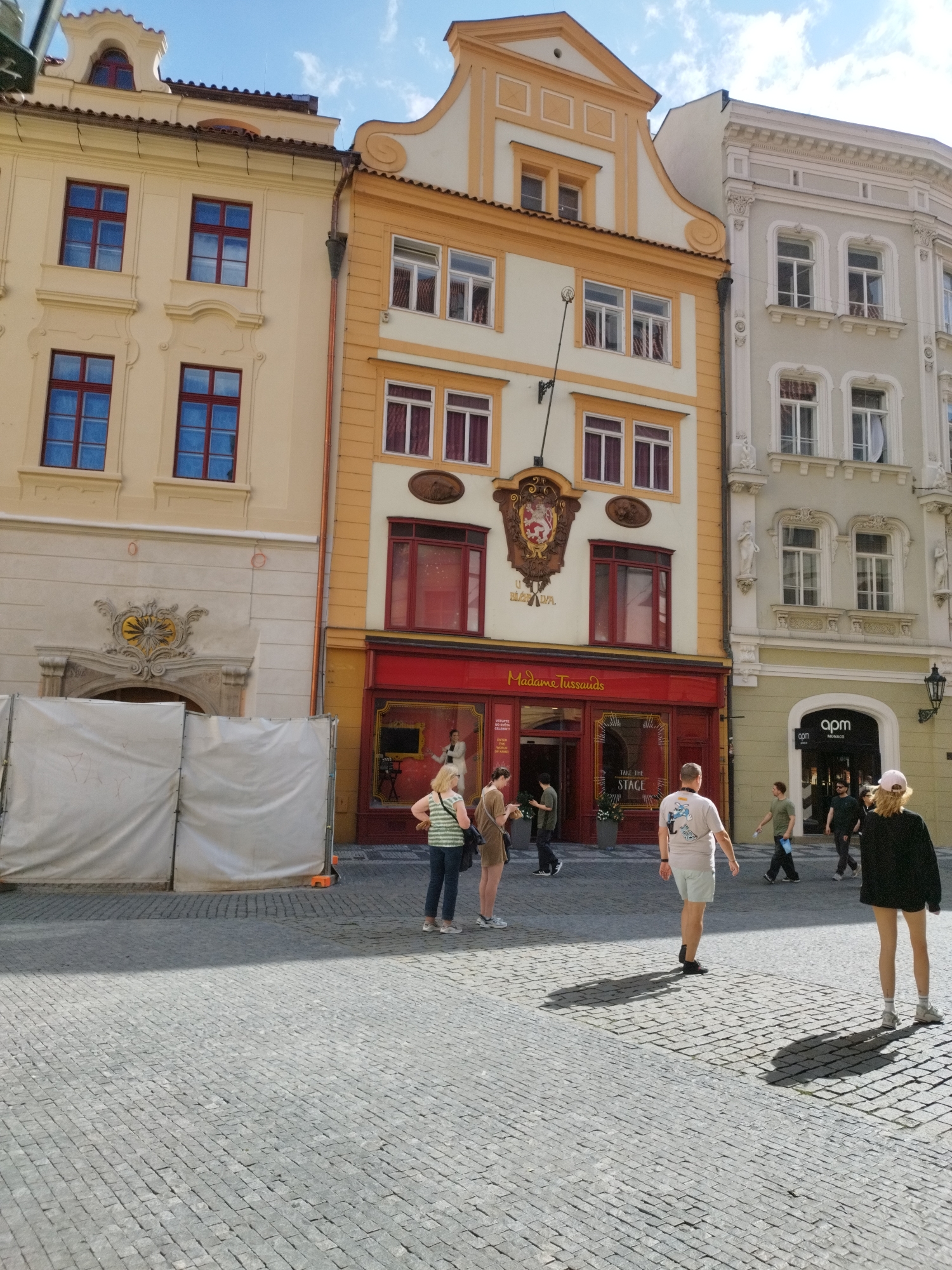 In the city of Unknown, Unknown, a bright, sunny day is depicted with a clear blue sky and scattered clouds. The scene is a cobblestone street in front of several buildings with ornate facades. The building on the left is cream-colored with red-trimmed windows. Next to it is a building painted in yellow and white, featuring a red-trimmed storefront labeled "Madame Tussauds." To the right of that is a light grey building with a curved entrance and a sign for "apm MONACO."
Several people are visible on the street. In the foreground, a person wearing a light-colored t-shirt and shorts walks away from the camera. To their left, a woman in a striped shirt and jeans stands with her hands on her hips, facing a doorway. Another person stands to her left, holding a bag. Further down the street, a man in a green t-shirt walks from right to left, and several other individuals are seen entering or near the "apm MONACO" store. A temporary white barrier is set up on the left side of the image, obscuring a portion of the cream-colored building's lower facade. Shadows are cast across the cobblestone pavement, indicating the direction of the sun.