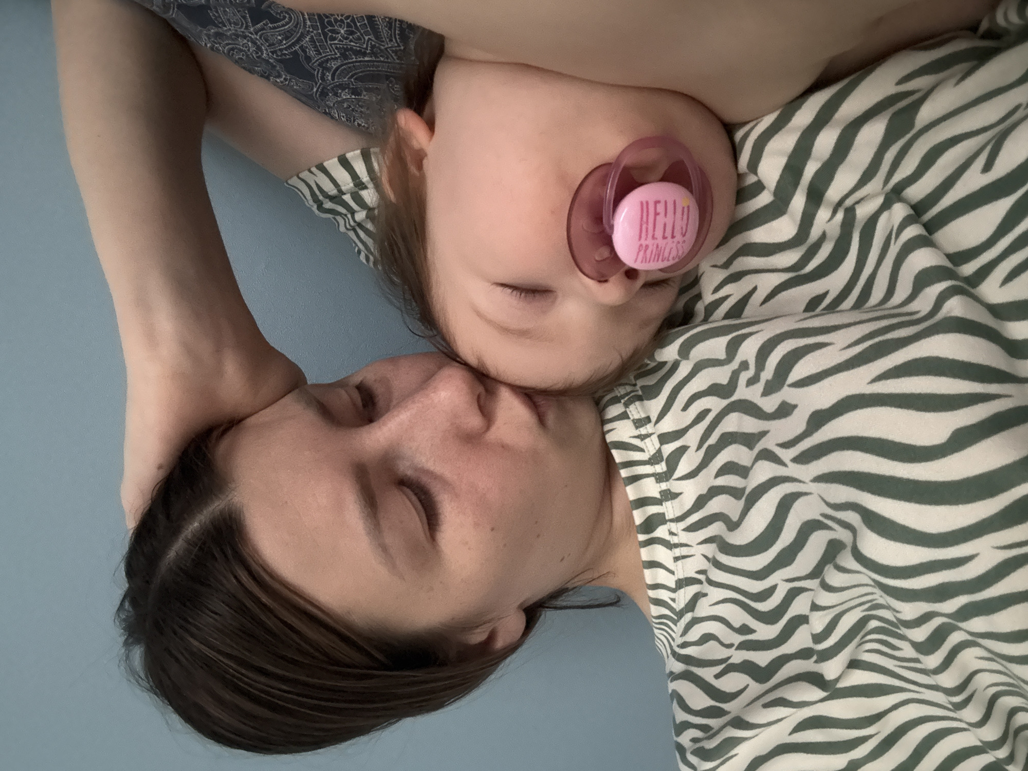 A woman and a young child are lying down on a blue surface. The woman has dark hair and her eyes are closed. The child, who is lying on the woman, has fair skin, closed eyes, and a pink pacifier in their mouth with the words "Hello Princess" on it. Both are wearing clothing with a green and white stripe pattern. The woman's hand is near her head. It is not possible to determine the location as Moscow, Russia, from the given visual information.