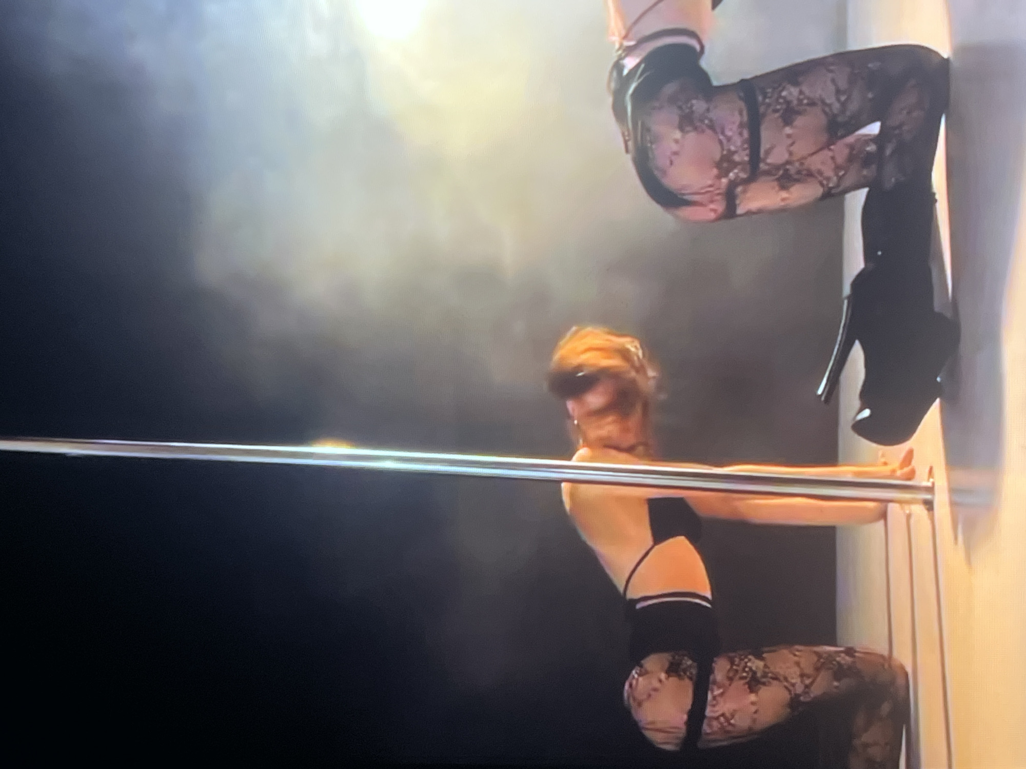 In a dimly lit performance space, two individuals are positioned on a polished floor next to a vertical pole. One individual, facing the viewer's left, is on all fours with their head lowered. They are wearing a black bikini-style top with white trim and black lace stockings. The other individual, to the right, is also on all fours, facing away from the viewer, their body arched. They are wearing a similar black bikini-style top with white trim and black lace stockings. A bright light illuminates the scene from the right, creating a hazy atmosphere. The background is dark, with some indistinct smoky or foggy elements. The floor appears to be a light-colored surface, possibly wood or laminate, reflecting the light. There is no indication of the city of Zoriane, Ukraine, in the image.