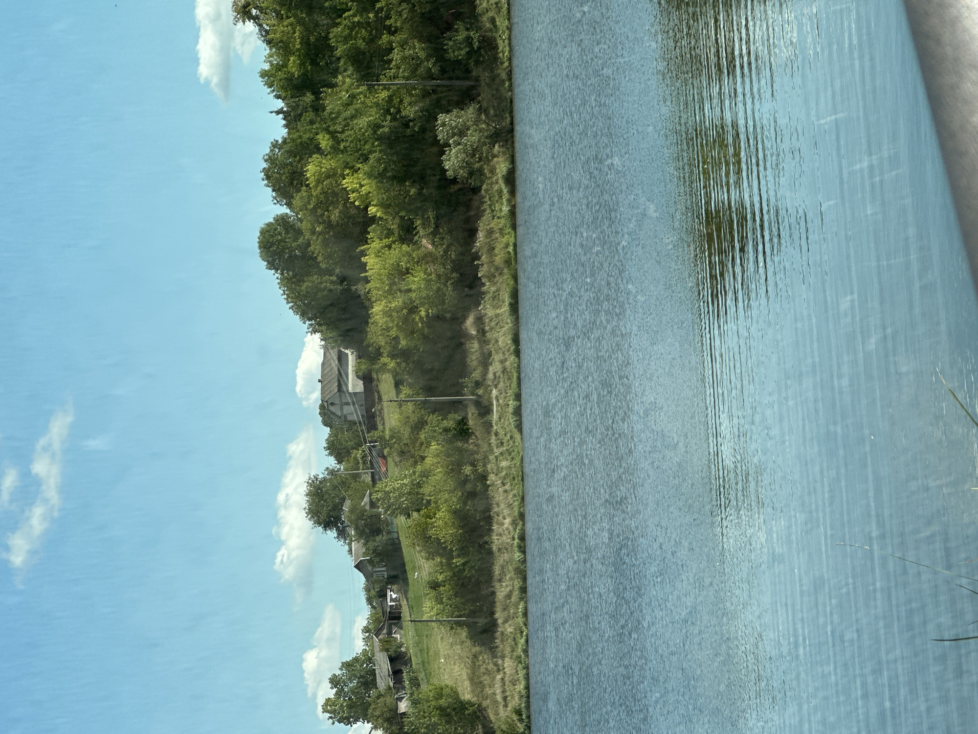 The image captures a tranquil scene in Makiv, Ukraine, featuring a wide expanse of blue water in the foreground. The water's surface is gently rippled, reflecting the bright blue sky with scattered white clouds above. Beyond the water, a lush green hillside rises, covered with a dense variety of trees and vegetation. Scattered across the hillside are several small, modest houses, suggesting a rural or semi-rural settlement. The houses have simple architectural designs, with pitched roofs and visible windows, appearing to be traditional village dwellings. A few utility poles are also visible along the slope. The overall impression is one of peaceful natural beauty, with human habitation integrated into the landscape.