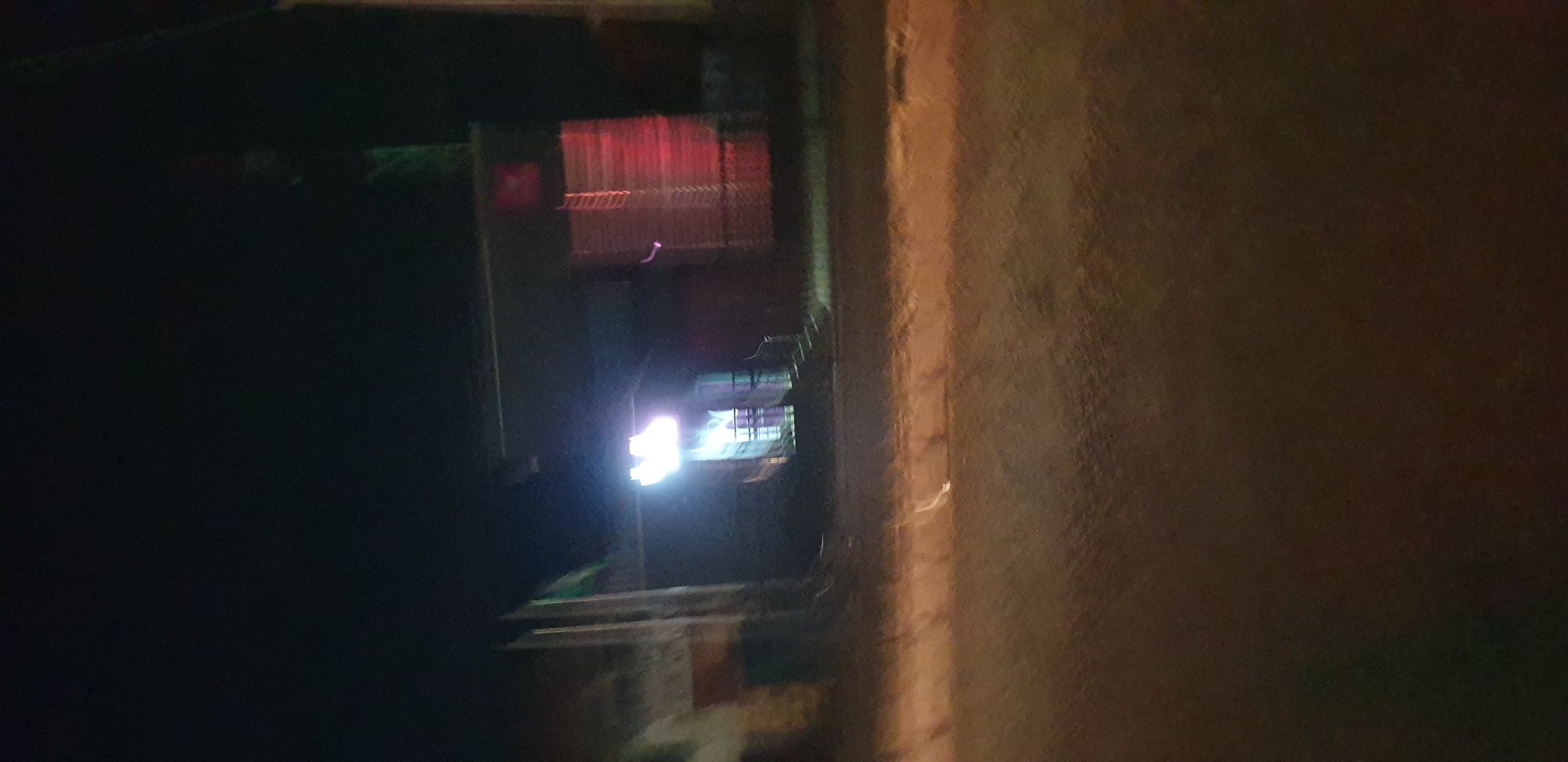 The image depicts a nighttime street scene in Sabon Tasha, Nigeria, characterized by extreme low light and significant motion blur. The foreground shows a dark, textured road or ground surface, faintly illuminated by ambient light. In the midground, to the left, a light-colored building with vertical features is vaguely discernible, accompanied by a pole displaying a partially unfurled green flag.
The central feature is a building entrance, intensely illuminated by a bright white or blue light source positioned above it, creating a strong glare that obscures details of the entrance itself, though steps leading up to it are faintly visible. To the right of this entrance, another structure or building exhibits a distinct reddish-orange glow, featuring vertical, repetitive patterns on its facade, possibly indicative of slatted or corrugated material. In the upper right background, a faint green structure, possibly a tower or antenna, is barely visible against the dark sky. No individuals or specific actions are discernible within the frame.