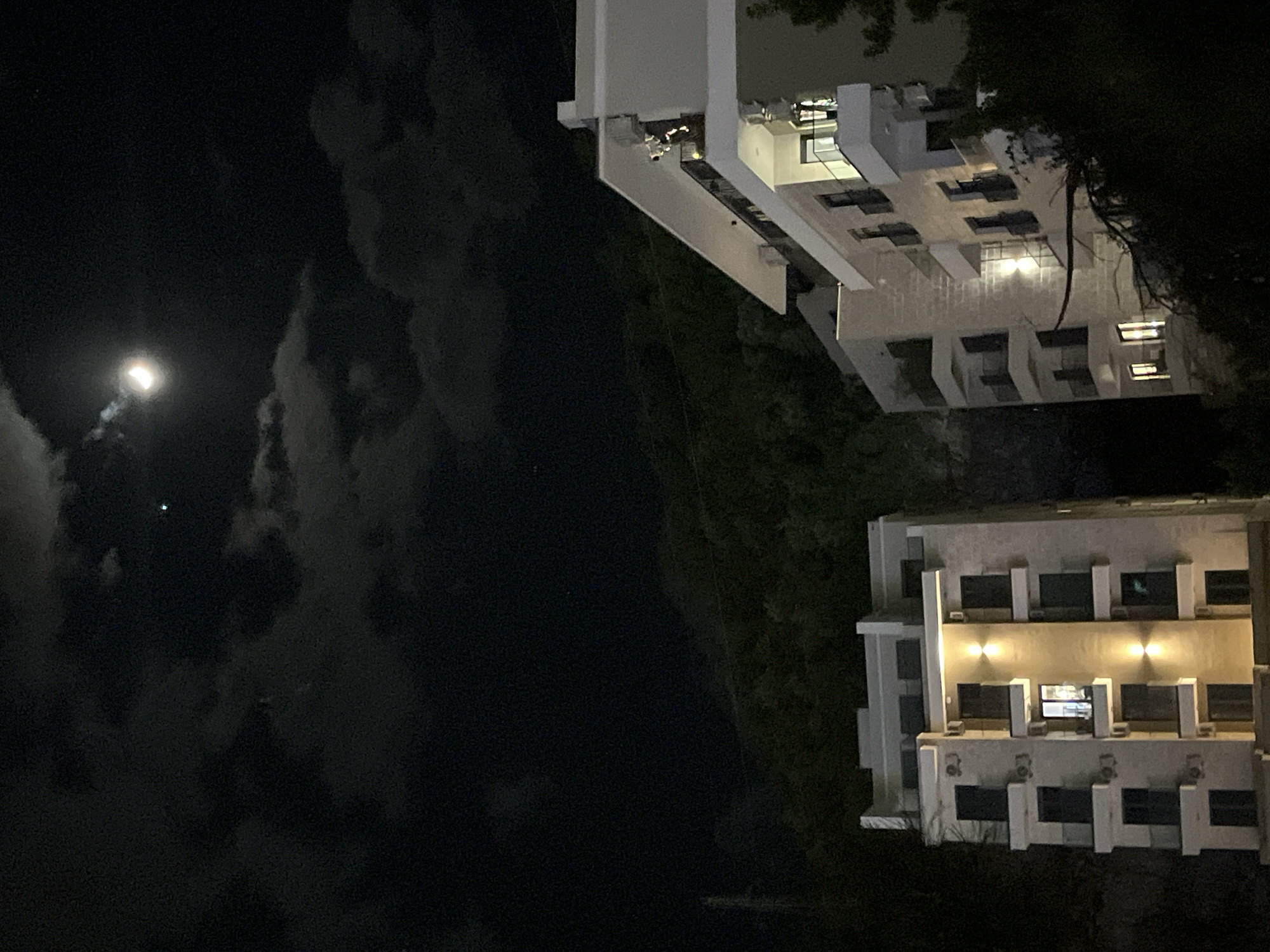 This nighttime image depicts a view of two modern buildings under a cloudy, moonlit sky. In the upper left quadrant, a bright, glowing moon is prominently featured, partially veiled by scattered clouds. A faint, tiny bright spot is visible above the moon, possibly a star or distant light source.
On the right side of the frame, two multi-story structures with light-colored facades are visible. The building closer to the viewer showcases a series of uniformly designed windows and balconies, with several exterior wall sconces emitting a warm yellow light. Some interior lights are also visible through the windows. Behind it, a second, similar building can be seen, also featuring contemporary architecture and illuminated interior spaces.
Dark, dense vegetation, likely trees or shrubs, forms a natural backdrop and outlines parts of the buildings, suggesting a lush, perhaps landscaped, environment. The scene is static, with no explicit human activity or event taking place. The overall impression is that of an urban or resort area at night, fitting the context of Budva, Montenegro. There is no visible text in the image.