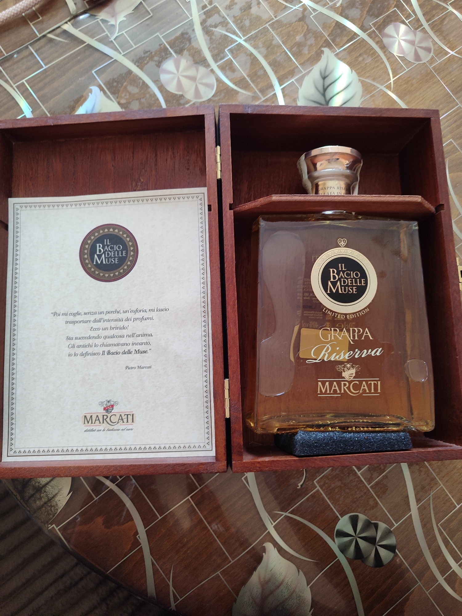 The image shows a wooden box containing a bottle of "Il Bacio Delle Muse Grappa Riserva Limited Edition" by Marcati. The bottle is placed on a small, dark gray foam block within the box. On the left side of the box is a paper insert with the Marcati logo and text in Italian describing the grappa. The background features a glossy, patterned surface with leaf and geometric designs. The scene's location is unknown, and it is not possible to discern any connection to Burgas, Bulgaria, from the image.