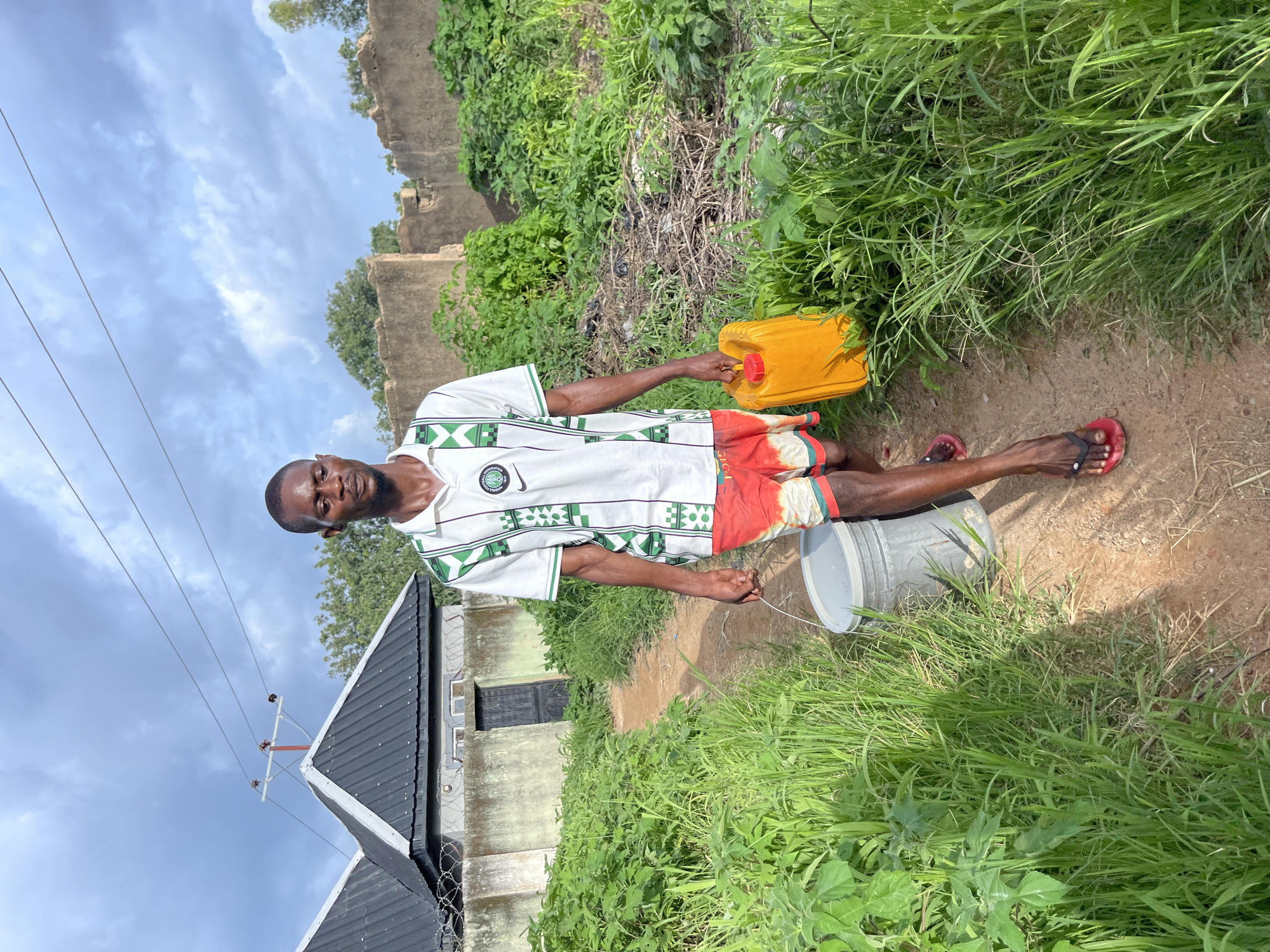 In Rafin Par, Nigeria, a young man walks on a dirt path towards the viewer, carrying a yellow jerrycan in his right hand and a gray bucket in his left. He wears a white and green patterned t-shirt, red and white patterned shorts, and red flip-flops. The path is bordered by tall green grass and lush vegetation. In the background, a building with a dark metal roof and a concrete wall stands to the left, while to the right, there is a dilapidated mud-brick structure and more dense greenery. Power lines are visible stretching across the cloudy sky above.