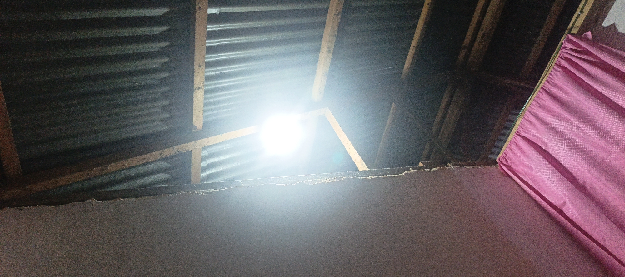 An interior shot of a dimly lit room with a bright light source dominating the center, appearing to be a bare bulb. The ceiling is constructed of corrugated metal sheets supported by wooden beams. To the right, a pink curtain is visible, partially obscuring the view of the ceiling structure. The foreground shows the top edge of a roughly textured wall. The image appears to be taken from below, looking upwards at the ceiling.