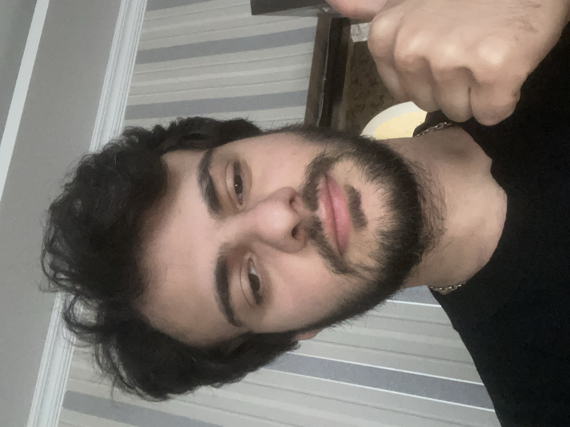 A close-up, selfie-style photograph shows a young man with dark, curly hair and a beard. He is wearing a black t-shirt and a silver chain. His right hand is raised, with his thumb up, indicating a "thumbs up" gesture. The background consists of a wall with vertical striped wallpaper in shades of gray and white, and a white crown molding along the top. The lighting suggests an indoor setting. There is no indication of the specific location within Tbilisi, Georgia.