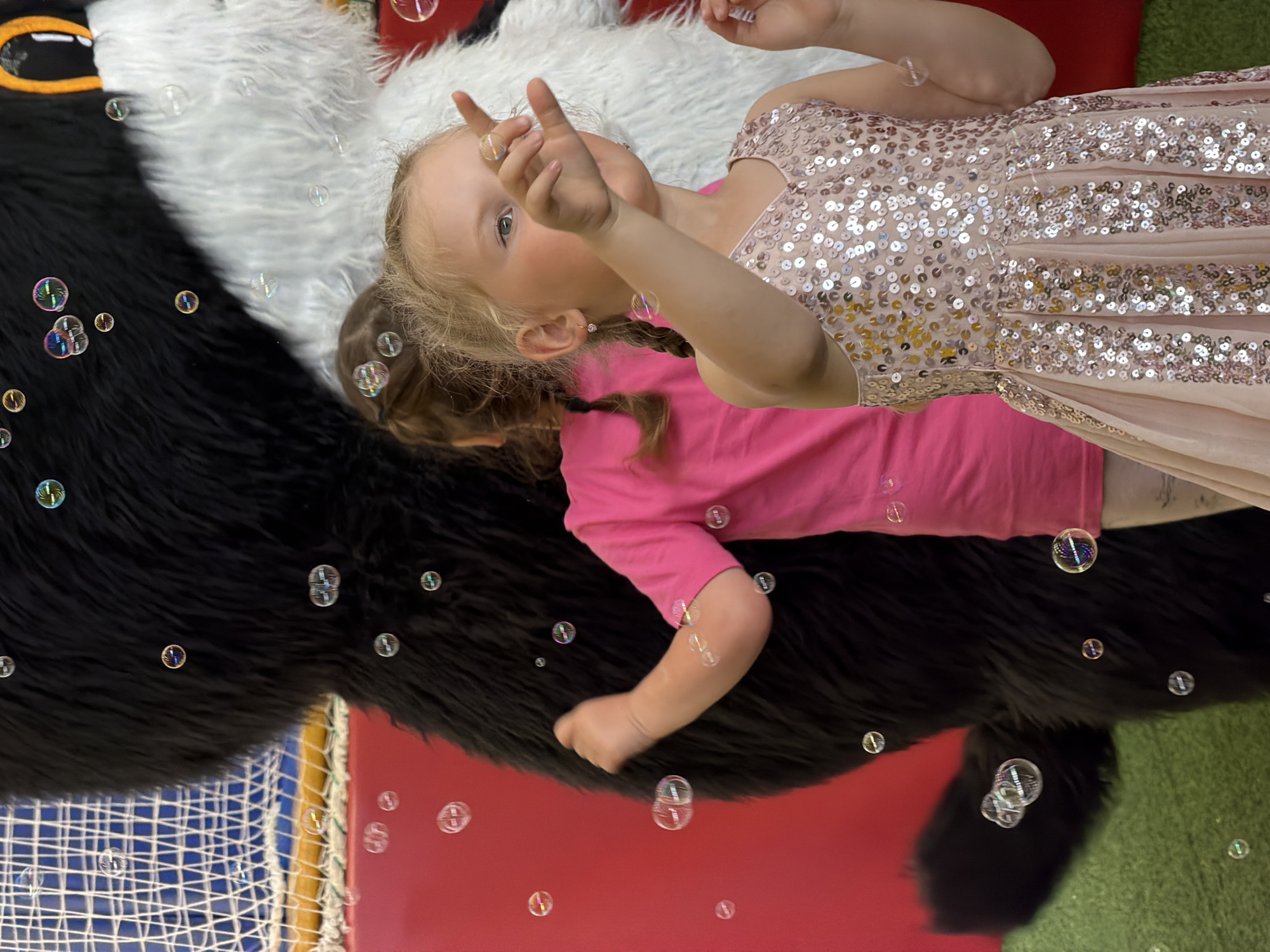 This image captures a lively scene with two young girls interacting with bubbles. The girl in the foreground, wearing a sparkling pink dress, is reaching out with her right hand, fingers extended, seemingly trying to touch or catch a bubble. Her eyes are focused on something beyond the frame, possibly the source of the bubbles. Behind her, another girl in a bright pink t-shirt is partially visible, her arm raised as if playing with the bubbles as well. They are standing in front of a large, furry mascot character, which is predominantly black with a white section on its right side. The mascot appears to be positioned in an indoor play area, suggested by a red wall and a green floor, possibly artificial turf. A white net structure is visible in the background to the left. The presence of numerous floating bubbles throughout the frame indicates that bubble blowing is actively occurring. This scene is likely taking place in an entertainment venue or a children's party.