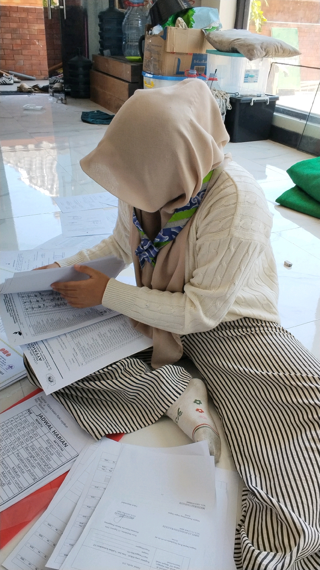 A person wearing a tan headscarf, a cream-colored cable-knit sweater, and a black and white striped skirt sits on a white tiled floor. The person is leaning forward and looking down at papers spread out on the floor in front of them. The papers appear to be documents or forms with text and tables. The person is holding one of the documents open with their left hand. A blue and green patterned scarf is visible around the person's neck. The background shows a room with a brick wall on the left, large plastic water jugs, stacks of boxes, and a window on the right. The lighting suggests it is daytime, with sunlight streaming in from the window. There are also some green cushions and other assorted items scattered around the floor in the background. The papers on the floor indicate this scene likely takes place in an office or educational setting. Given the provided context, the scene is located in the city of Mojokerto, Indonesia.