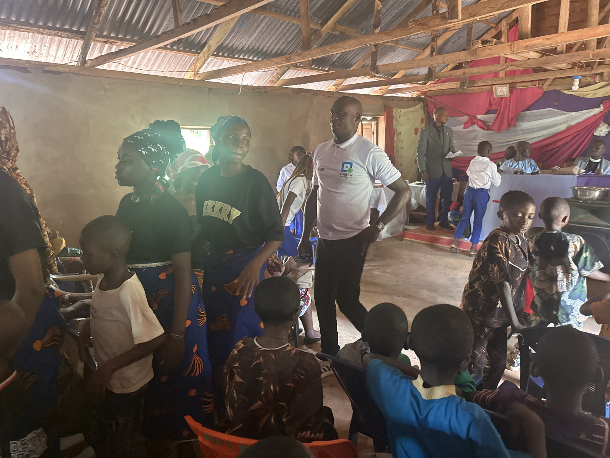 Inside a rustic structure with a corrugated metal roof and concrete walls, several people are congregated. The setting appears to be a simple hall or meeting space. The atmosphere seems casual, with individuals both standing and seated.
A man in a white polo shirt with the word "GREEN" and another word below it is standing with his hands on his hips, partially obscured by children in the foreground. Several women and children are visible, wearing a variety of clothing.
At the back of the room, there's a slightly elevated platform with people seated behind a table covered in cloth. A man in a dark suit is standing near this platform, possibly addressing the group. The table backdrop is decorated with drapes of various colors.
The lighting in the room is dim, with some light streaming in from outside. The presence of seating and the elevated platform suggests a meeting or event is taking place.