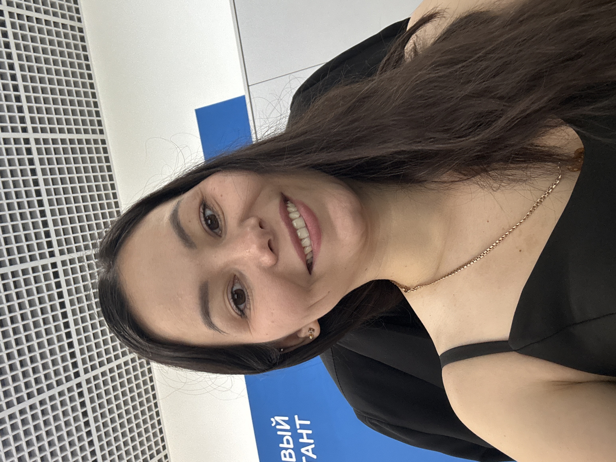 A woman with long, dark hair and brown eyes smiles at the camera in a close-up selfie. She is wearing a black dress with thin straps and a gold chain necklace. Her left ear has two piercings, one with a small stud and another with a dangling earring. Behind her, a blue sign with white Cyrillic text is partially visible, and a white tiled ceiling is overhead. The scene appears to be indoors, possibly an office or public building, with a hint of grey paneling on the right side. The text on the blue sign, "ВЫЙ ГАНТ," suggests a location possibly related to business or services. The city of Vavilovo, Russia, is not discernible from the visual elements in the image.