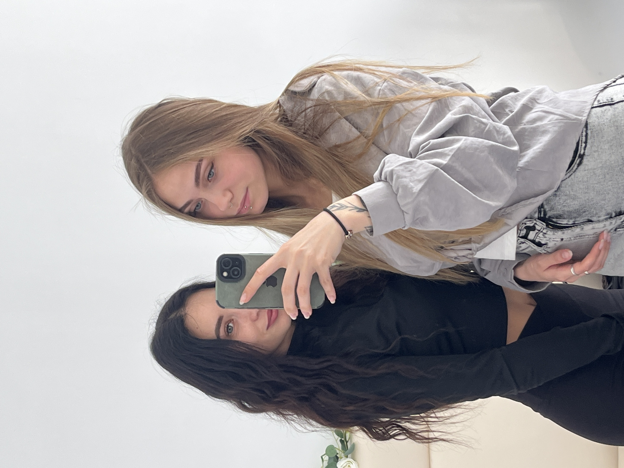 Two women are posing for a selfie. The woman on top has long blonde hair and is wearing a light gray button-down shirt and acid wash jeans. The woman on the bottom has long dark hair and is wearing a black long-sleeved shirt. They are both holding a green phone. The background appears to be a white wall. The location is Фоки, Russia.