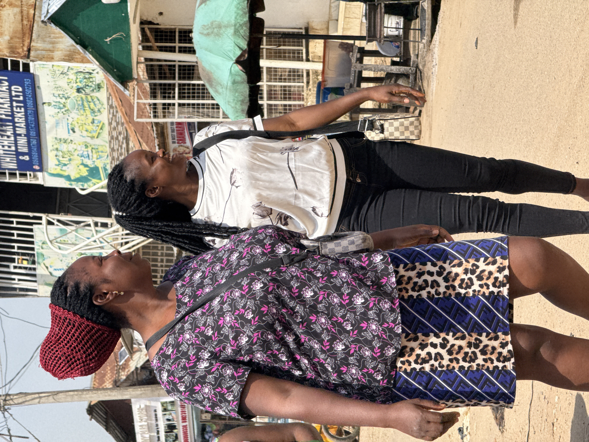 In the city of Abuja, Nigeria, two women are walking outdoors on a sunny day. The woman on the left is wearing a red knitted head covering, a black and pink floral print short-sleeved shirt, and a blue skirt with a leopard print and diagonal white stripes. She has a black bag with a checkered pattern slung across her body. The woman on the right is wearing a white t-shirt with a subtle floral pattern and black skinny jeans. She has black braided hair. They are walking on a dirt and asphalt road. In the background, there are buildings, one with a blue sign for "WHITEHEART PHARMACY & MINI-MARKET LTD". There are also market stalls and an open umbrella. The overall scene depicts a typical street view in a bustling Nigerian city.