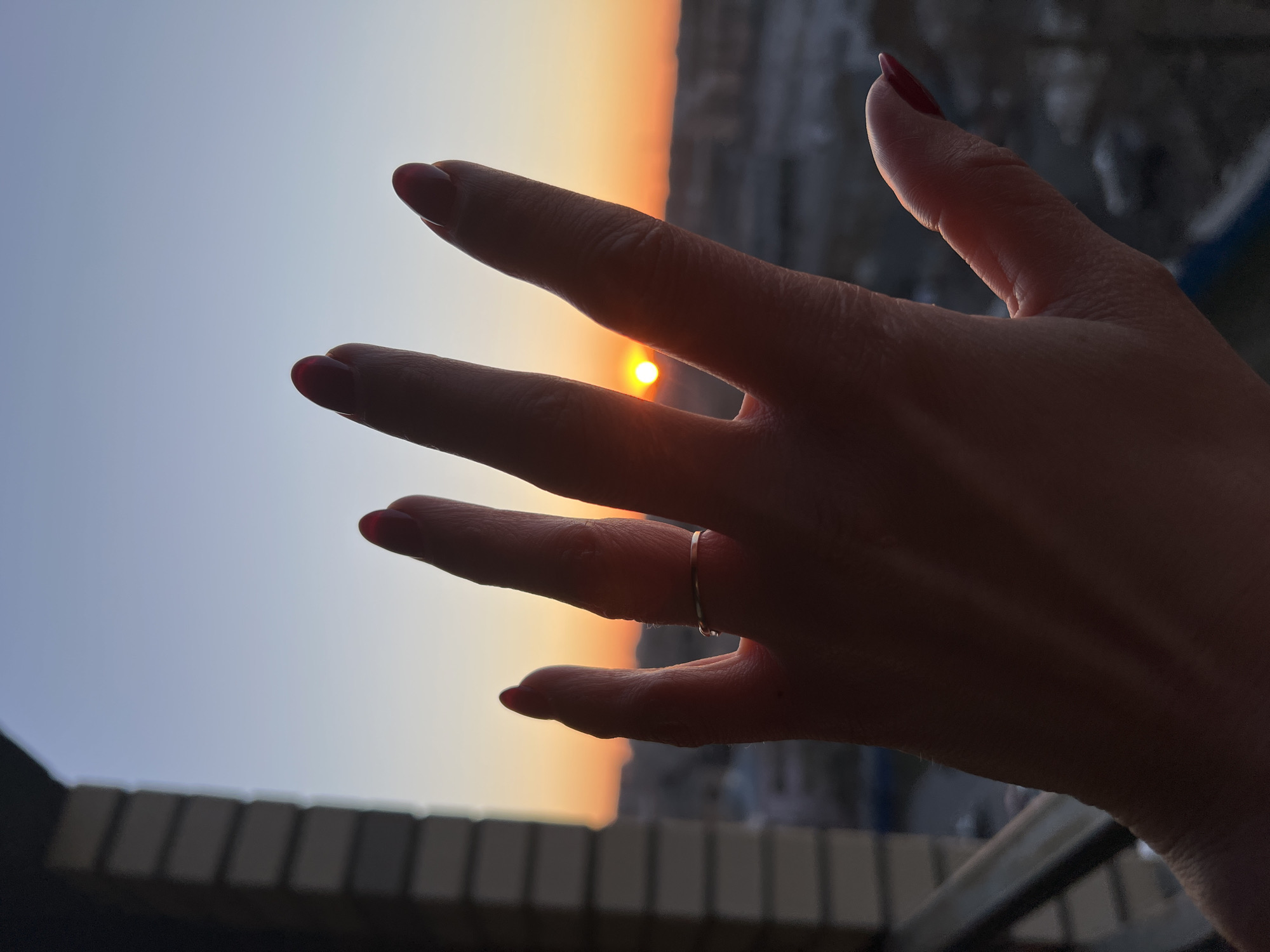 A right hand, adorned with a simple silver ring on the ring finger and red nail polish on all fingernails, is positioned in front of a sunset in what is suspected to be Saint Petersburg, Russia, judging by the architectural style of the buildings visible in the background. The sun appears to be setting between the index and middle fingers. The horizon is visible between the fingers. The lower portion of the frame shows a section of a building facade or balcony with regular horizontal lines.