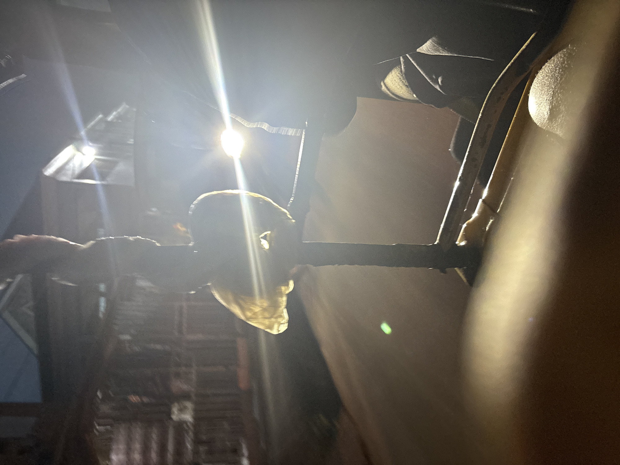 The image is a nighttime street scene in Onitsha, Nigeria. The foreground is dominated by a blurry yellow object, possibly part of a vehicle's interior. Behind this, a dark metal pole with a yellowish, translucent material wrapped around its upper section is visible. Bright headlights from a vehicle create lens flares and illuminate the scene, suggesting movement or stationary activity on a road. In the background, a wooden fence is visible, with faint lights and building structures suggesting a residential or commercial area. The overall lighting is dim, with strong highlights from artificial light sources. The image appears to be taken from within a vehicle, looking outwards.
