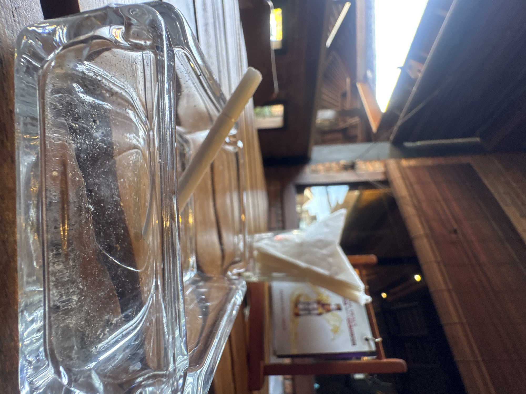 An ashtray with a cigarette resting in it is in the foreground of the image. The ashtray is made of clear glass and sits on a wooden table. In the background, a stack of white napkins in a glass holder is visible, along with a sign advertising "Special Deals." The overall setting appears to be an outdoor dining area with wooden beams and a thatched roof, suggesting it is likely a restaurant or cafe. The location is Kuta Selatan, Indonesia.