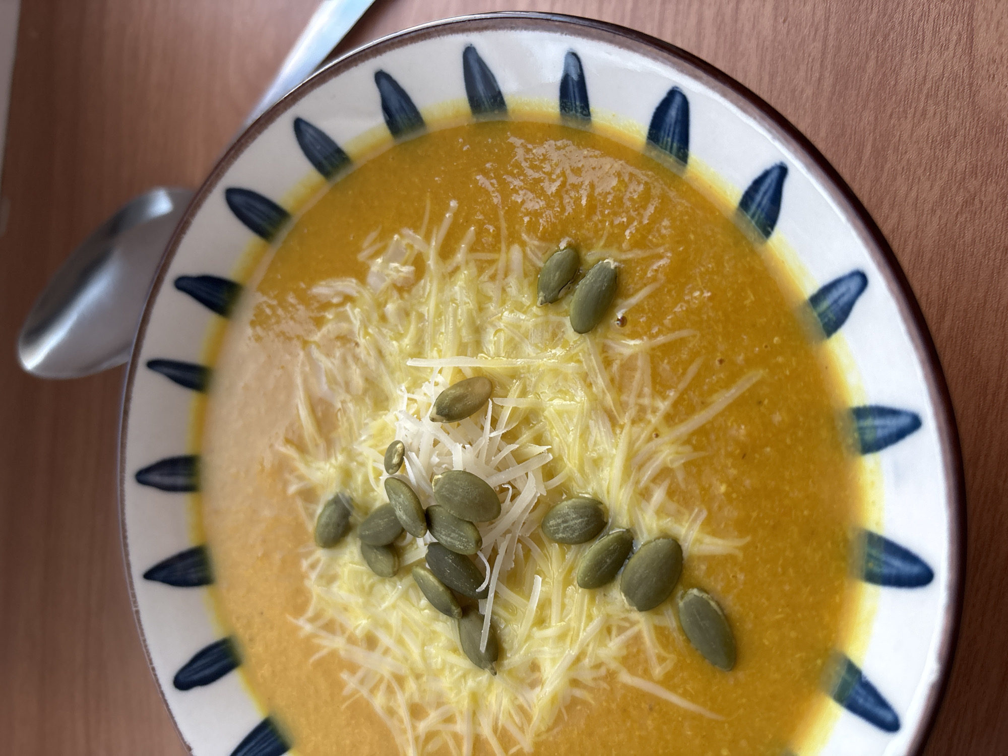 A top-down shot depicts a bowl of pumpkin soup with a garnish of shredded cheese and pumpkin seeds, placed on a wooden surface. The bowl has a white base with a blue, triangular pattern around the rim. A spoon rests beside the bowl. The location is not discernible in the image, thus I cannot say if it is in Perm, Russia.